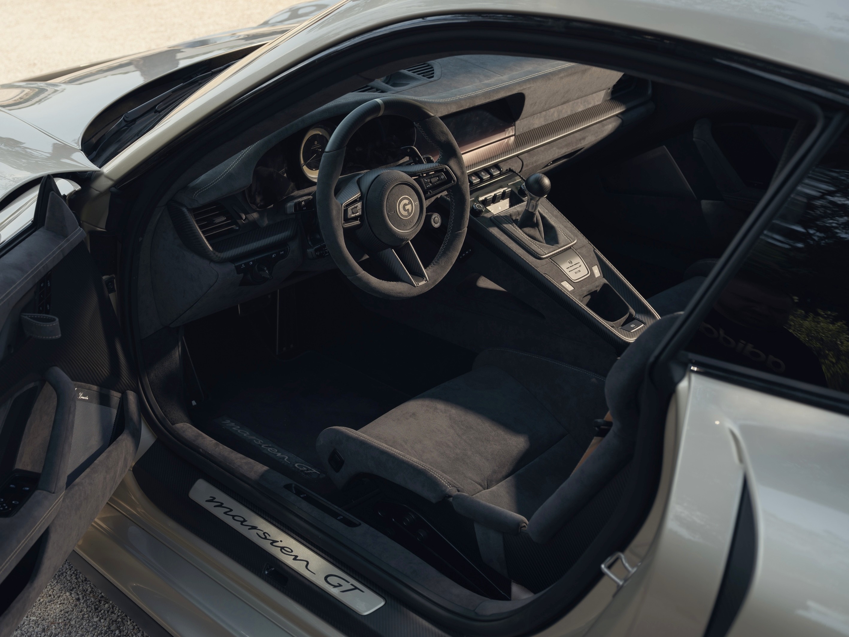 Interior de um carro esportivo cinza com a porta aberta, mostrando o volante, painel, câmbio manual e banco do motorista em suede escuro. A soleira da porta exibe a inscrição marsien GT