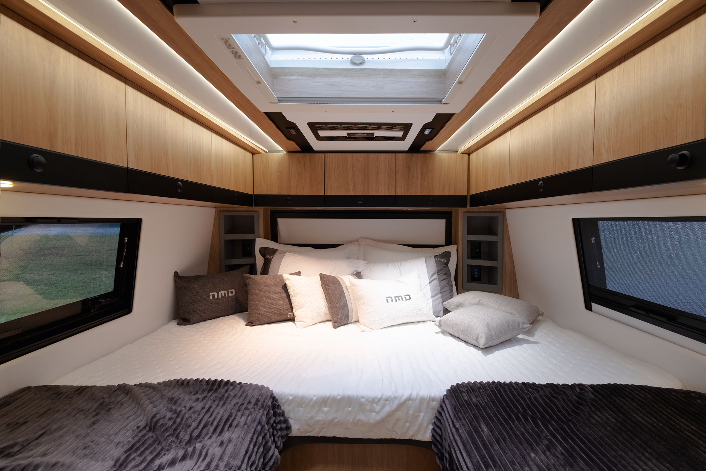 Interior de um quarto de motorhome com cama de casal central, lençóis brancos, travesseiros cinzas e brancos com a inscrição nmd, e cobertores cinza escuro nas laterais. Armários de madeira clara com iluminação embutida e uma claraboia no teto. Janelas retangulares escuras nas laterais mostram a paisagem verde externa