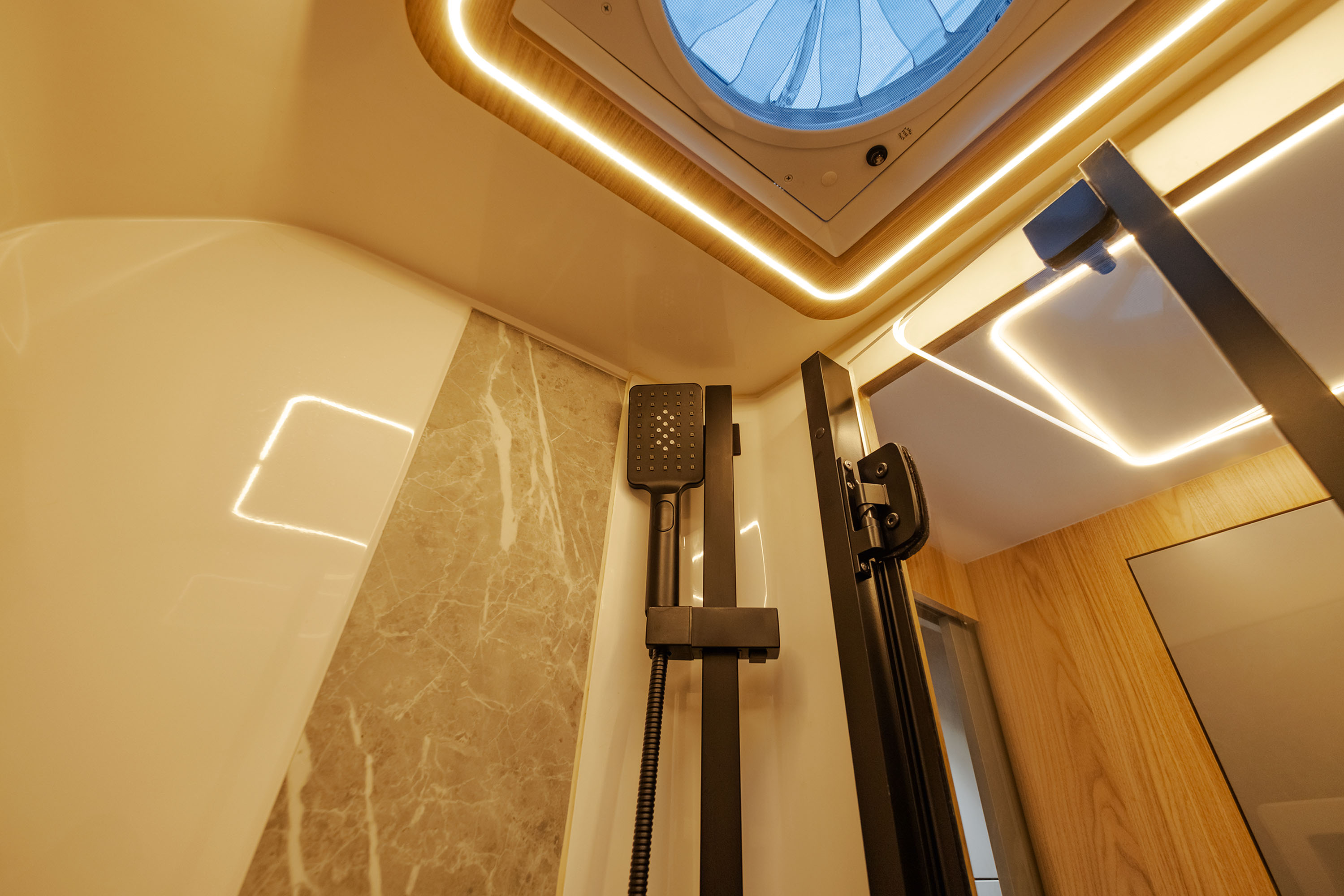 Interior de um banheiro moderno com chuveiro preto fosco, paredes claras e detalhes em mármore. O teto possui uma claraboia redonda com ventilador e iluminação LED embutida, criando um ambiente sofisticado e funcional