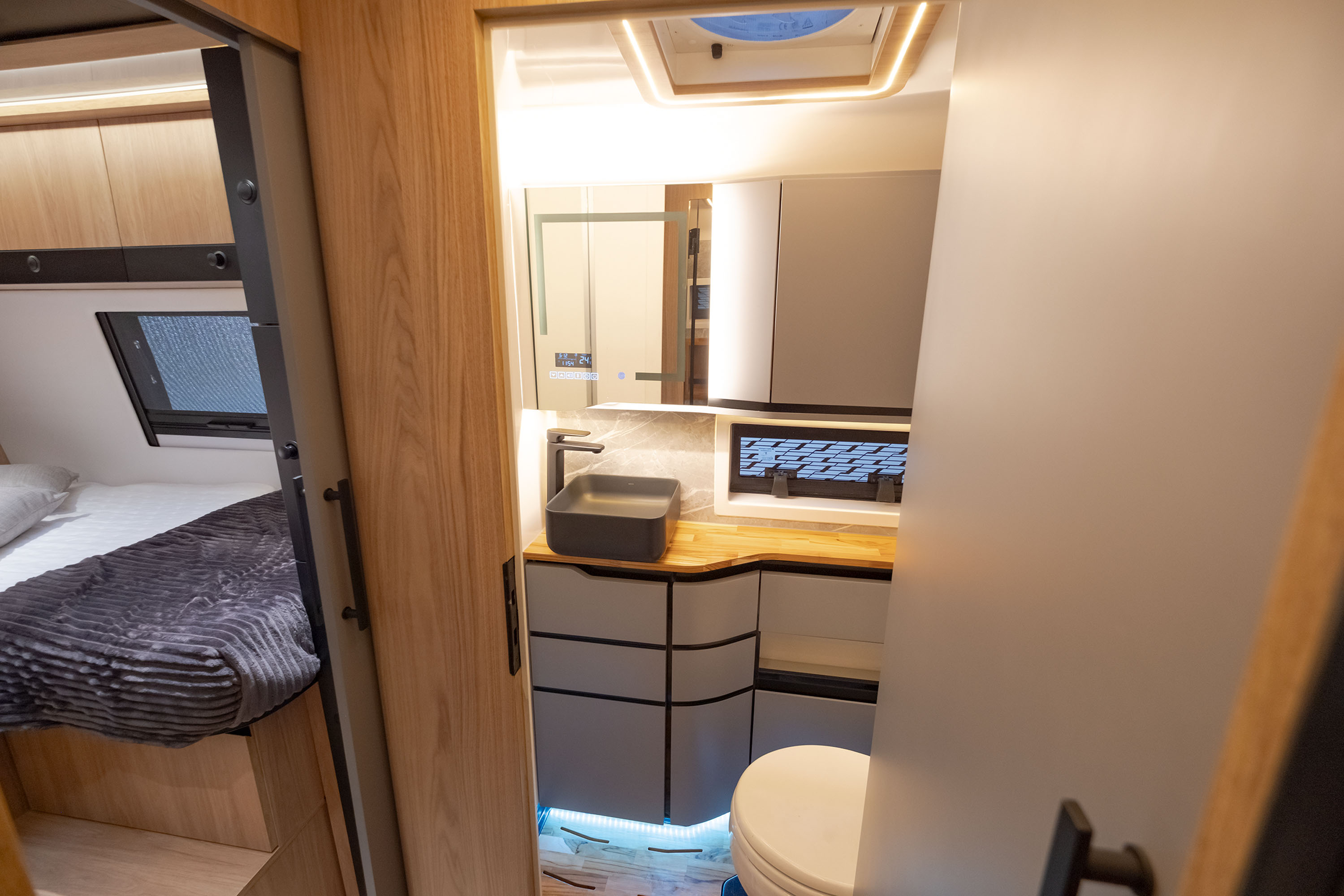 Interior de um motorhome moderno, mostrando um quarto com cama e um banheiro compacto. O quarto tem uma cama com lençóis brancos e uma manta cinza escura, com armários de madeira clara acima. O banheiro, à direita, possui uma pia retangular cinza escura sobre um balcão de madeira, armários cinzas e um vaso sanitário branco. Há iluminação embutida no teto e sob os armários do banheiro, criando um ambiente acolhedor