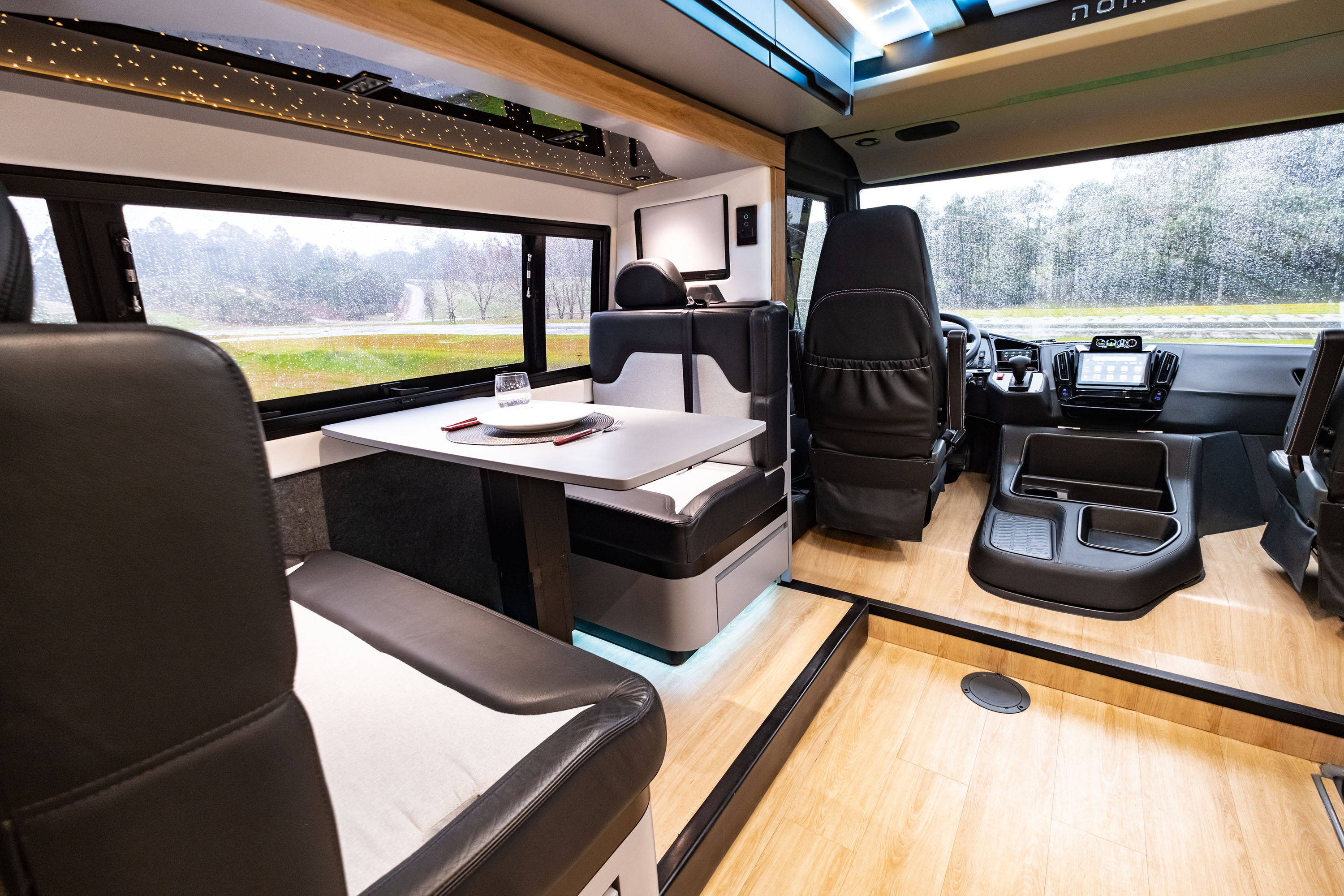 Interior de um motorhome moderno com piso de madeira clara, bancos estofados em preto e branco, e uma mesa com prato e copo. Ao fundo, janelas mostram uma paisagem verde com árvores e uma estrada, e o painel de controle do veículo.