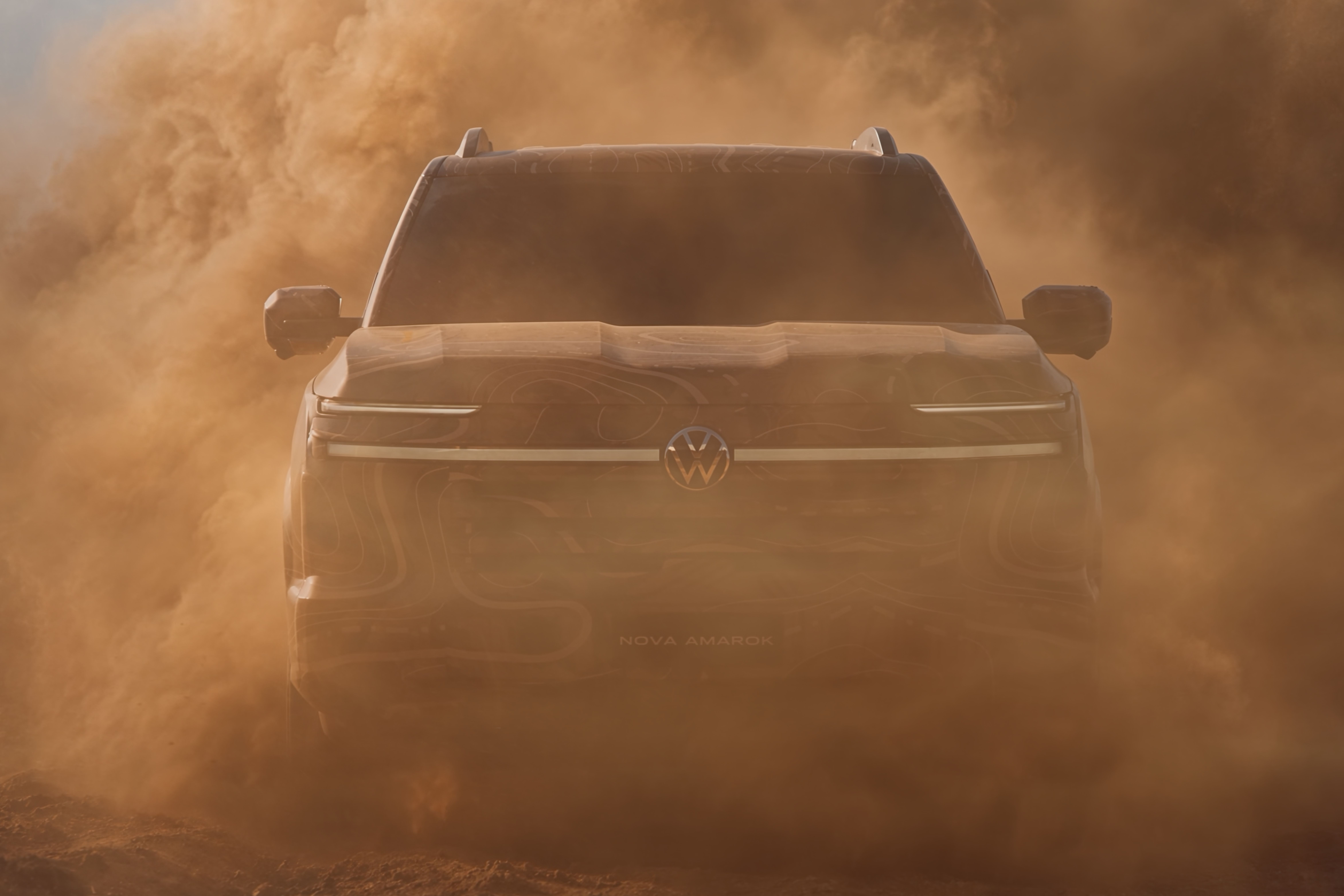 Picape Volkswagen Amarok escura, parcialmente visível em meio a uma nuvem de poeira marrom-alaranjada, com o logo VW na grade frontal e a inscrição NOVA AMAROK abaixo