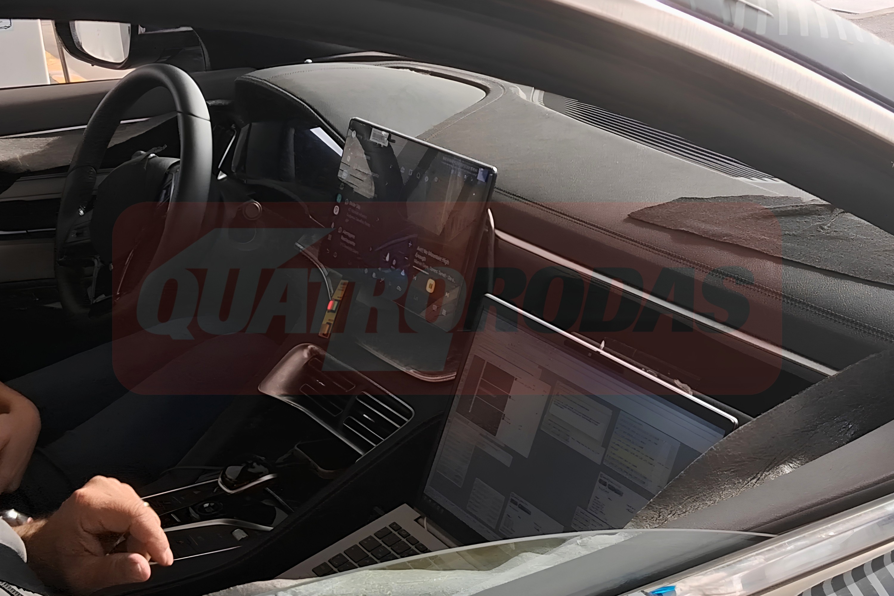 Interior de um carro com volante à esquerda, painel escuro e um tablet vertical no centro. Um notebook prateado está aberto no banco do passageiro, refletindo a luz. Uma mão masculina aparece no canto inferior esquerdo. A marca dágua QUATRO RODAS está sobreposta