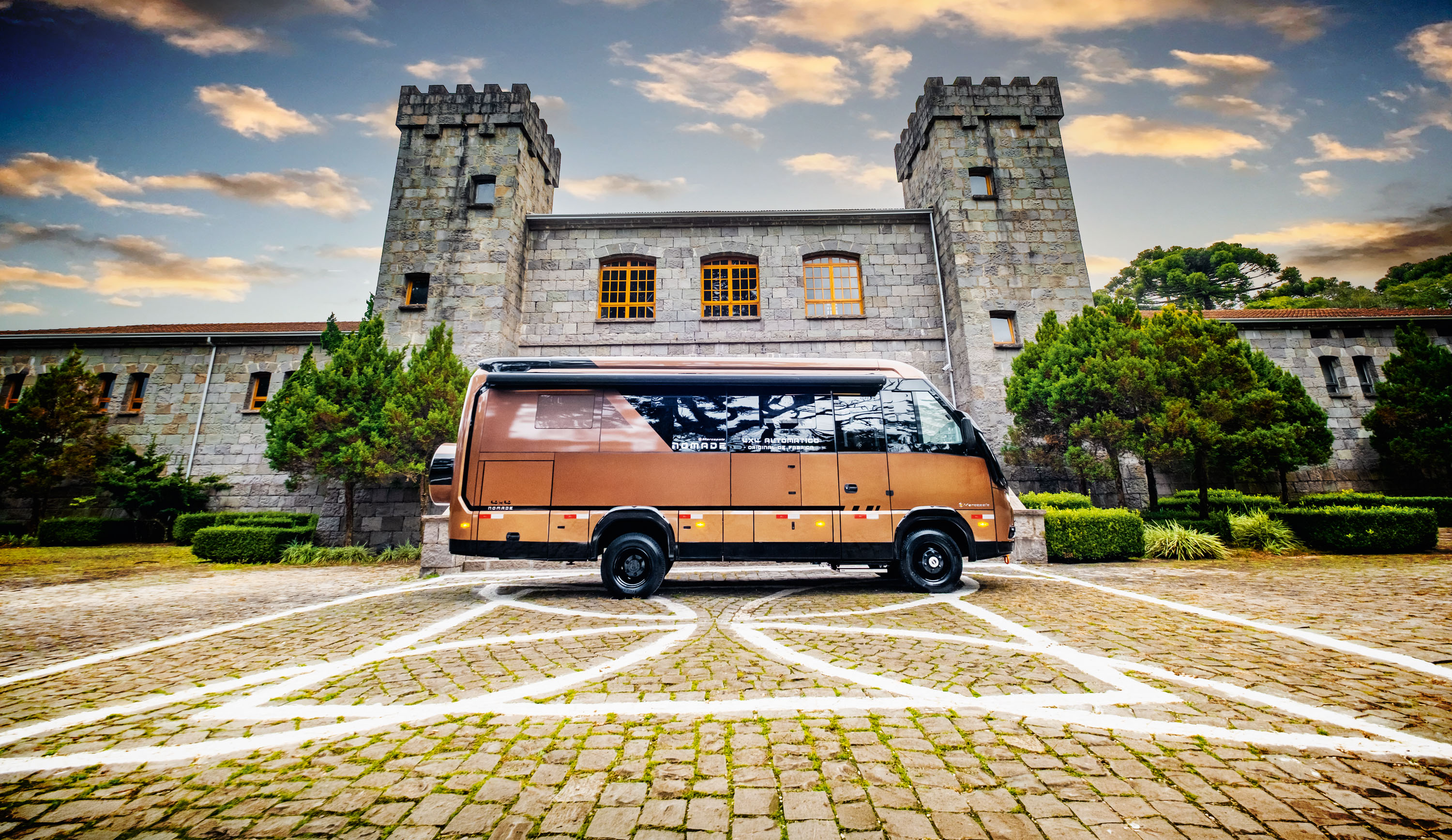 Motorhome marrom-bronzeado estacionado em p&aacute;tio de paralelep&iacute;pedos com marca&ccedil;&otilde;es brancas, em frente a um edif&iacute;cio de pedra com duas torres e janelas amarelas, sob c&eacute;u azul com nuvens