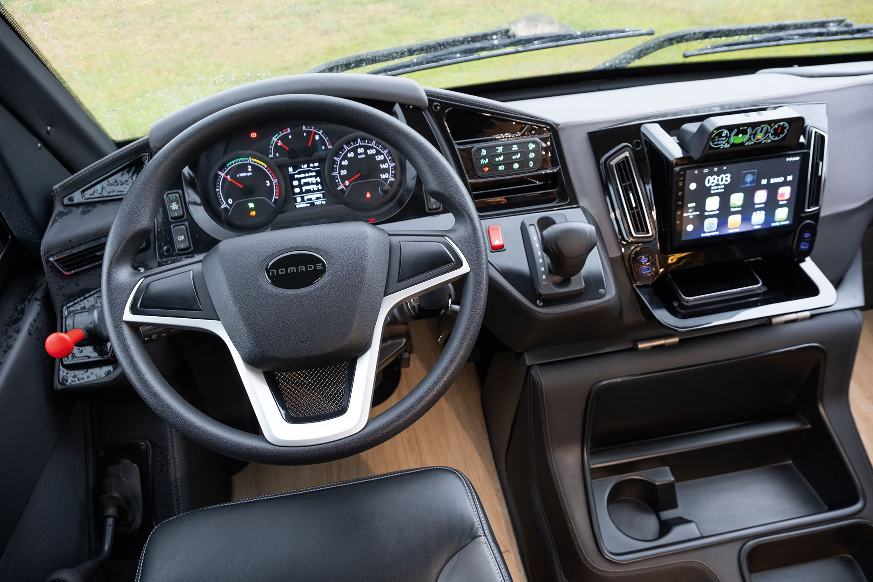 Interior de um veículo com volante preto e detalhes prateados, painel de instrumentos digital, tela multimídia com ícones de aplicativos e um smartphone em um compartimento abaixo. À direita, câmbio automático e porta-copos.