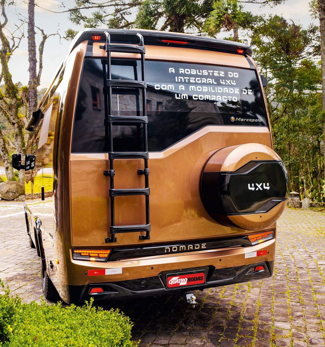 Traseira de um motorhome Marcopolo Nomade 4x4 cor bronze, com escada preta, estepe e a frase A robustez do integral 4x4 com a mobilidade de um compacto no vidro. Estacionado em pátio de pedras, com arbustos e árvores altas ao redor