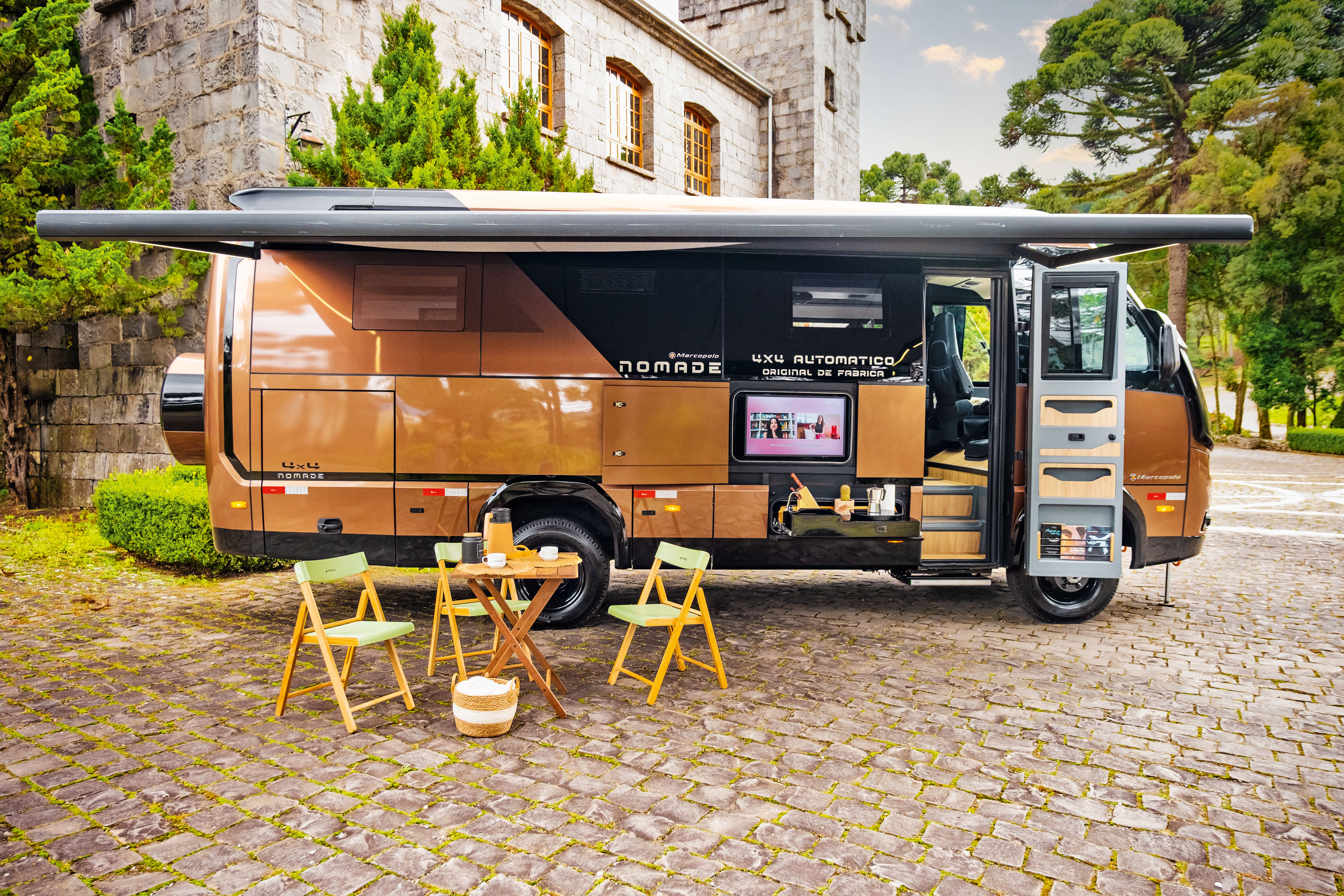 Motorhome Nomade marrom e preto estacionado em pátio de pedras, com toldo aberto e mesa de madeira com cadeiras dobráveis à frente. A lateral do veículo exibe uma TV e compartimentos abertos, revelando utensílios de cozinha. Ao fundo, um edifício de pedra com janelas e árvores verdes