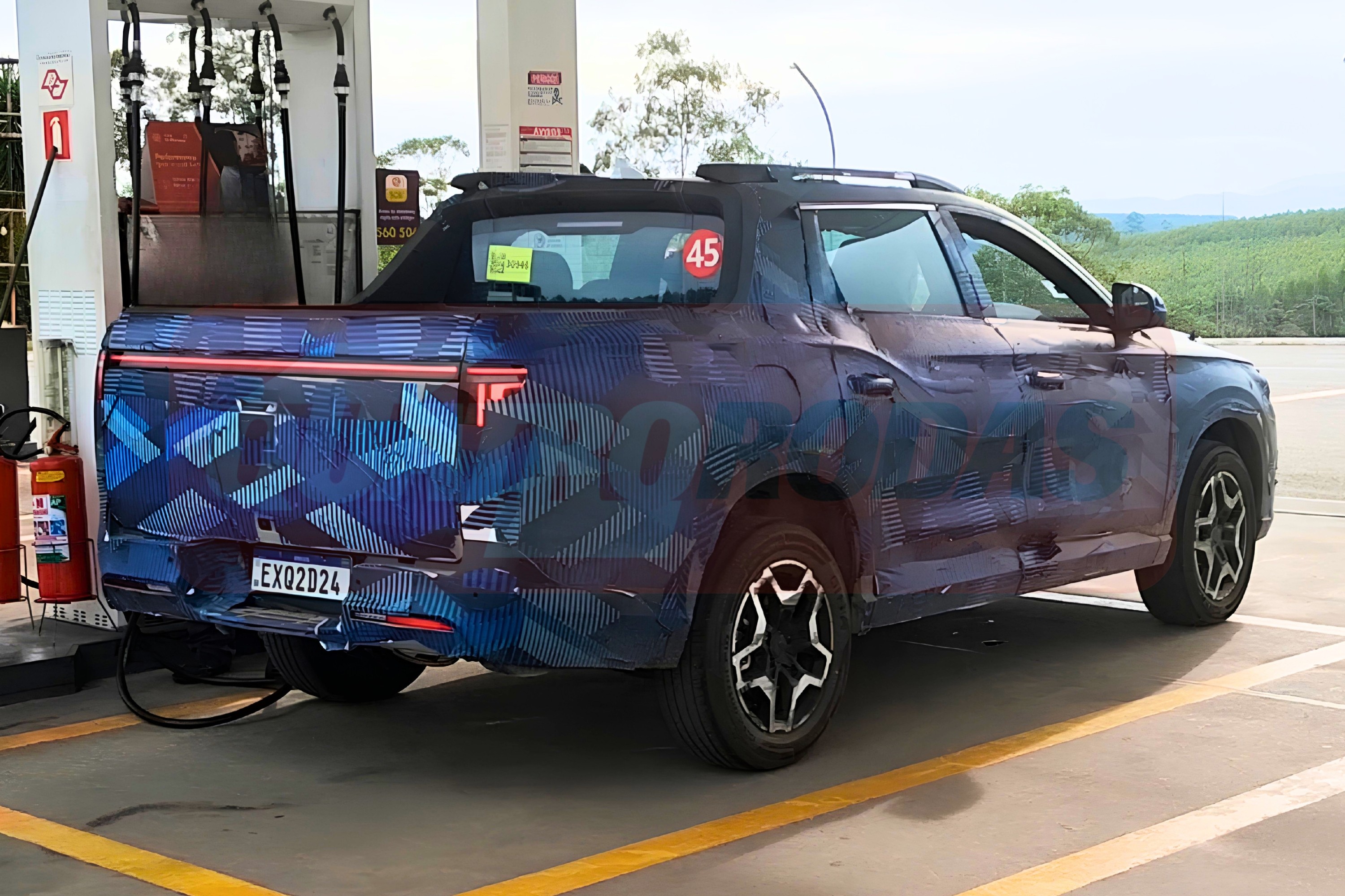 Picape camuflada azul e preta, com padr&atilde;o geom&eacute;trico, estacionada em posto de gasolina, vista traseira lateral. A placa &eacute; EXQ2D24 e h&aacute; um adesivo vermelho com 45 na janela traseira. Ao fundo, &aacute;rvores e c&eacute;u claro