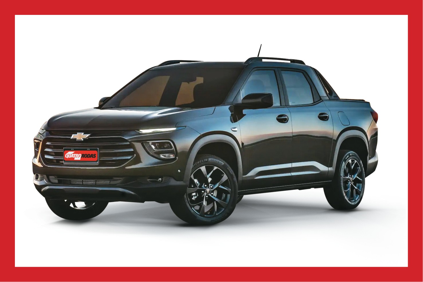 Picape Chevrolet Montana cinza escura, vista de três quartos frontal, com faróis acesos, grade preta e placa vermelha Quatro Rodas na frente. Possui rack de teto e rodas esportivas pretas, destacando-se em fundo branco