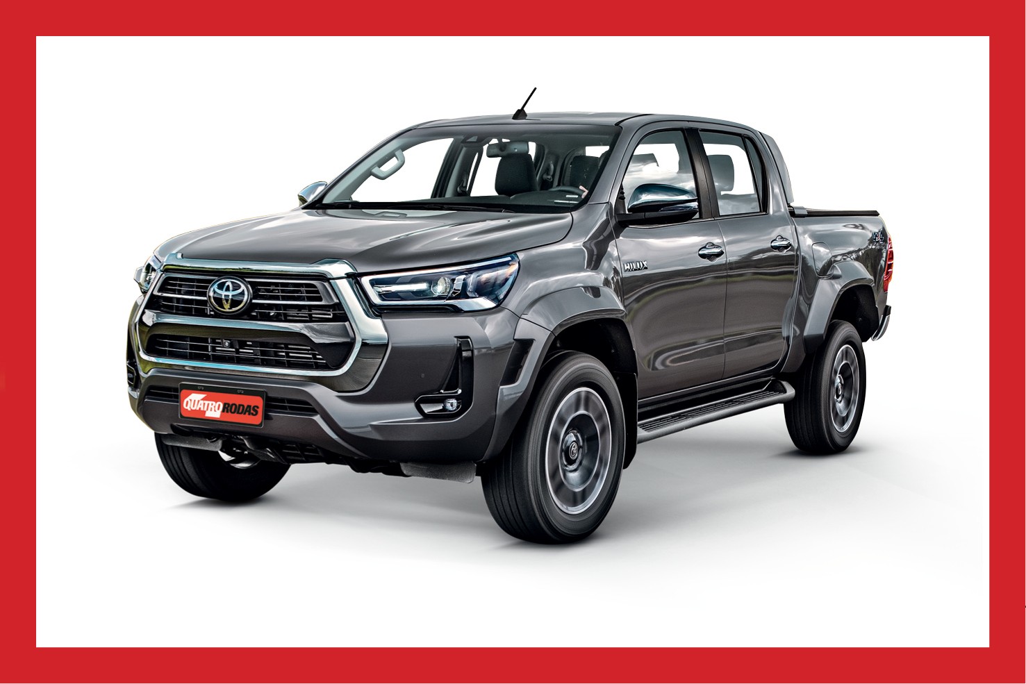 Picape Toyota Hilux cinza escura, vista de frente e lateralmente, com a placa QUATRO RODAS na parte inferior da grade, sobre fundo branco