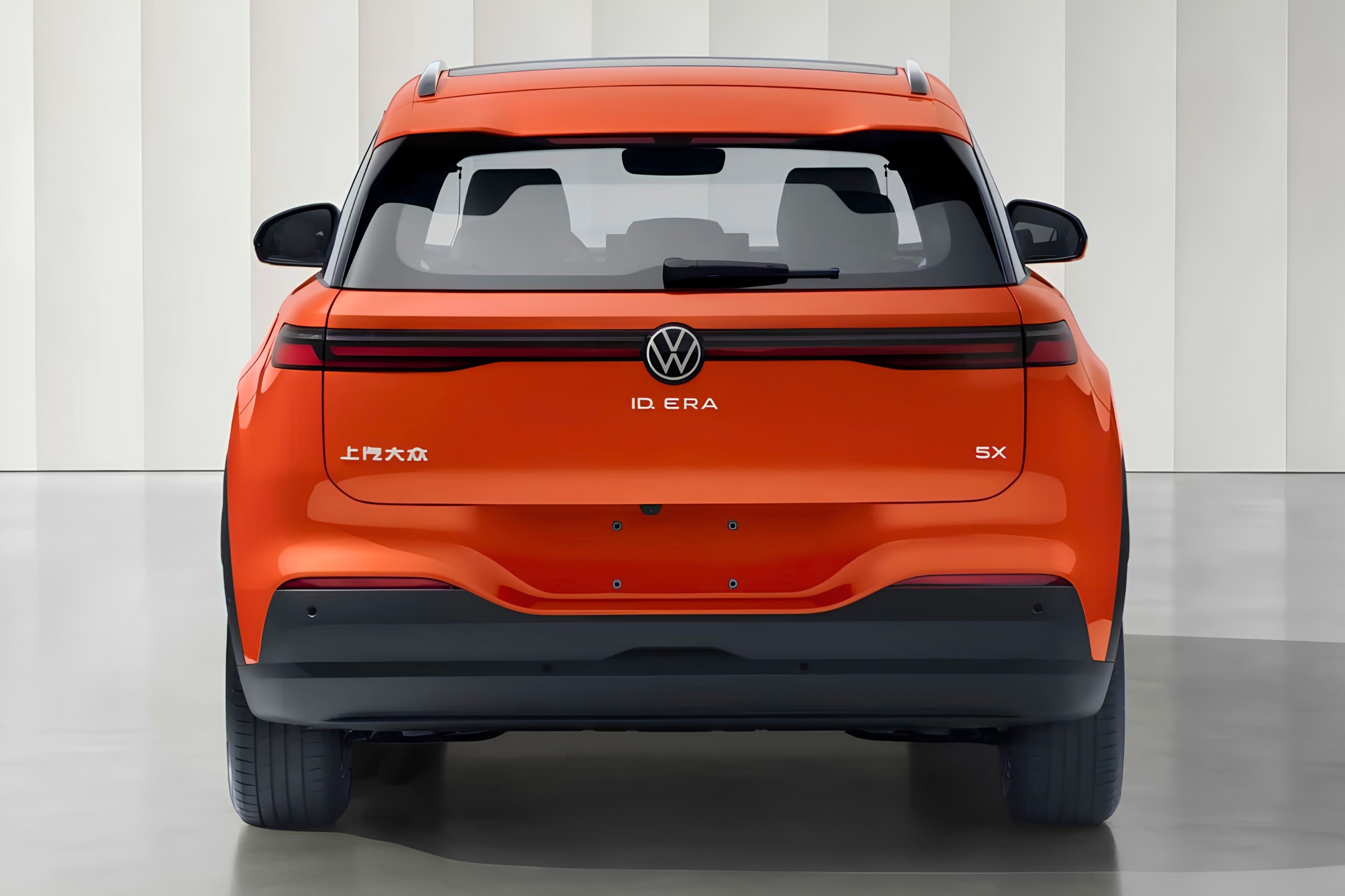 Traseira de um SUV elétrico Volkswagen ID.ERA laranja, com o logo VW e o nome do modelo centralizados. À esquerda, caracteres chineses e à direita, 5X. O carro está em um estúdio com paredes brancas e piso cinza