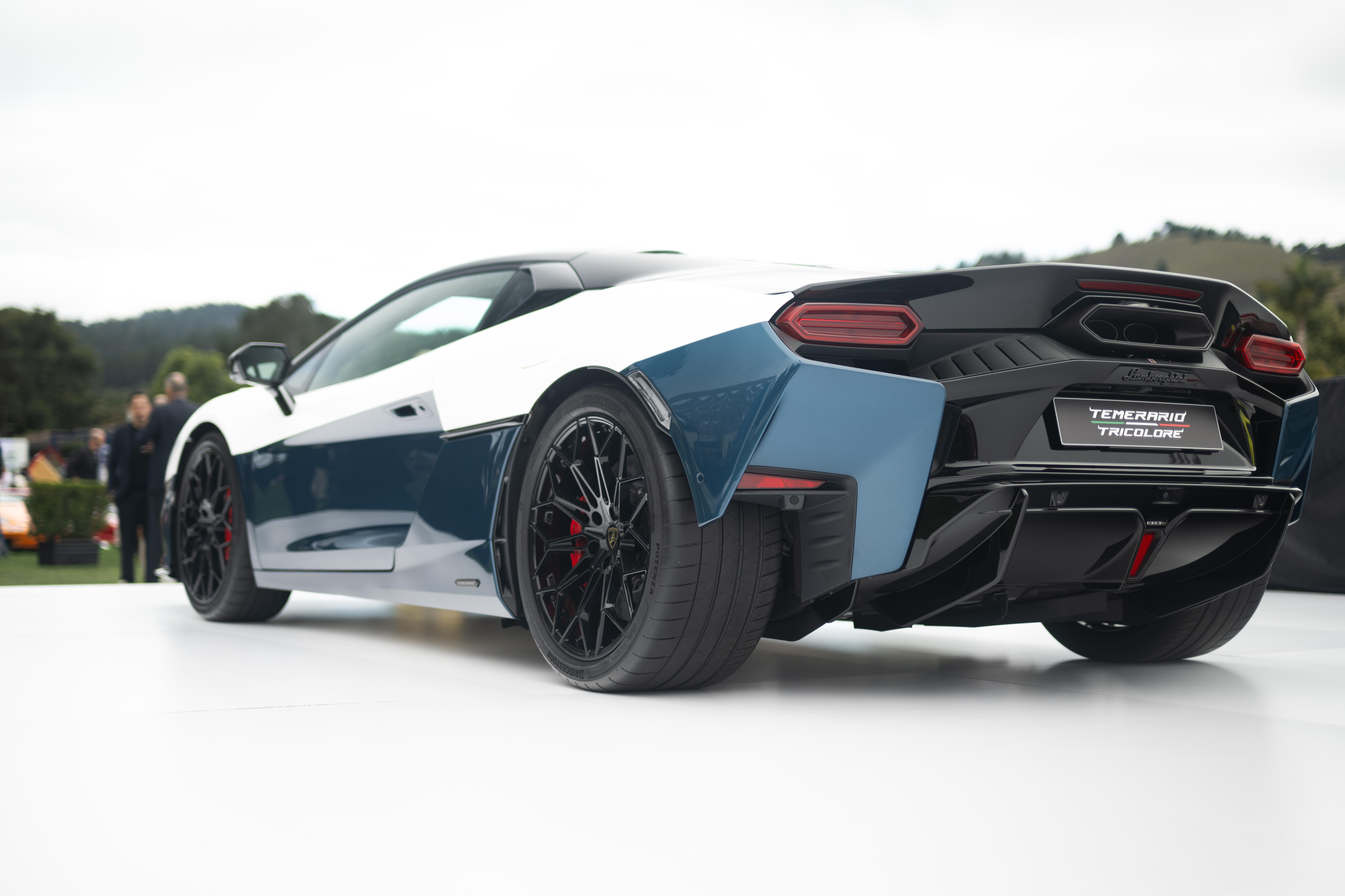 Carro esportivo branco e azul-marinho, visto de trás, com rodas pretas e pinças de freio vermelhas. O para-choque traseiro é preto com difusores e a placa exibe TEMERARIO TRICOLORE. Pessoas desfocadas e vegetação ao fundo, sob céu claro