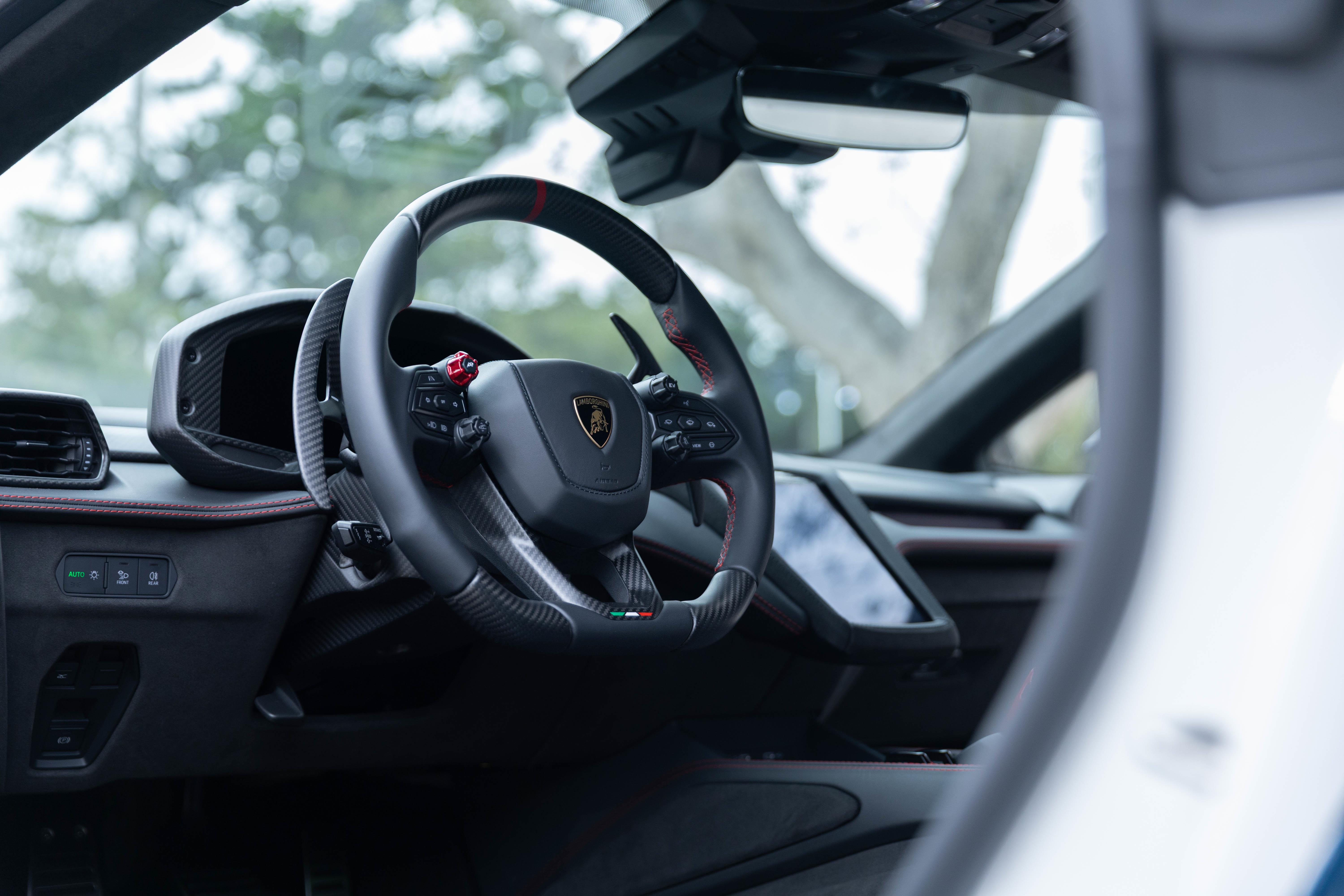 Interior de um carro esportivo de luxo, com volante preto de couro e fibra de carbono, detalhes em vermelho e o emblema da Lamborghini ao centro. Ao fundo, o painel digital e a tela multimídia, com a paisagem externa desfocada através do para-brisa