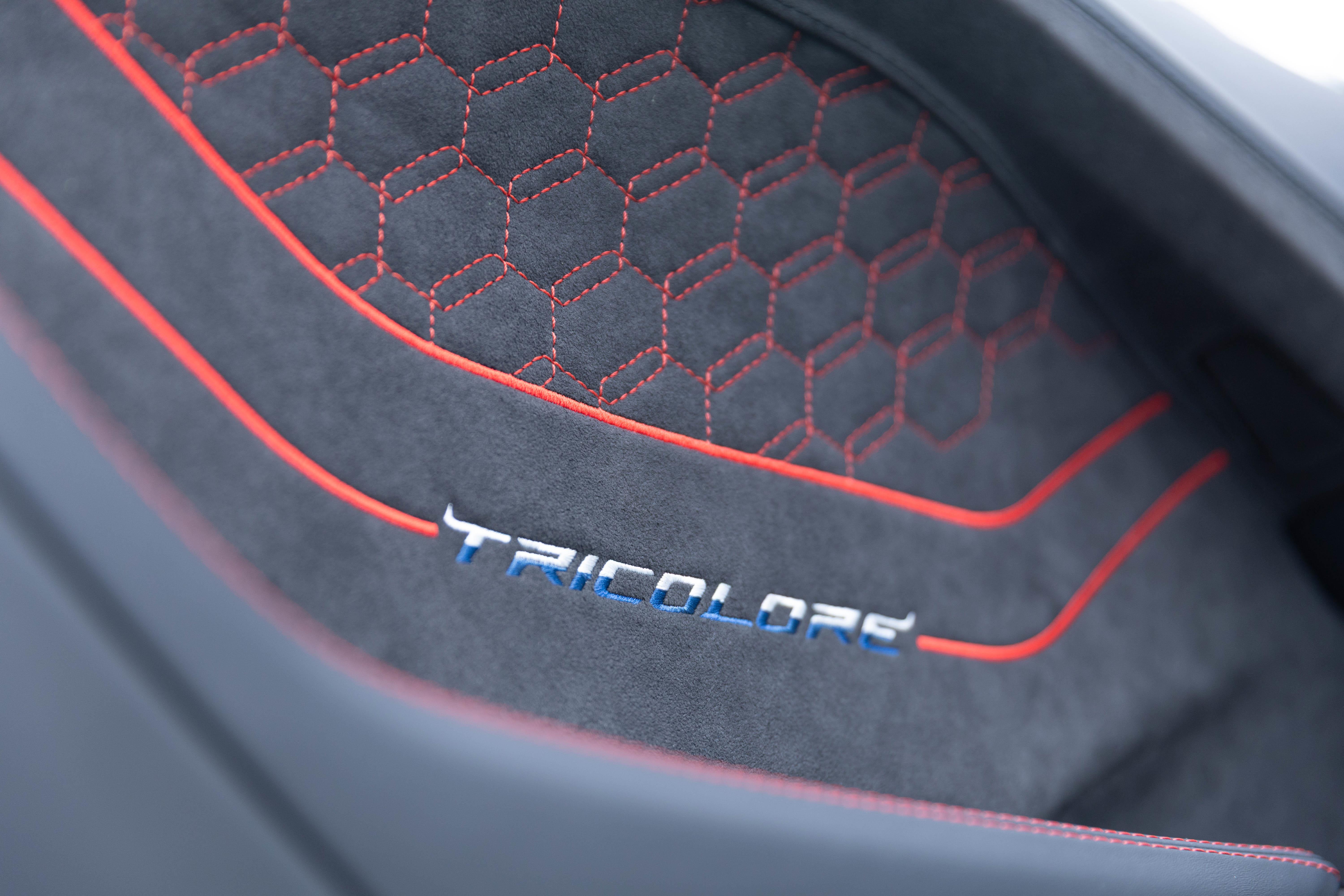 Detalhe do estofamento de um carro, em tecido cinza escuro com costuras vermelhas em padrão hexagonal e duas linhas vermelhas curvas. A palavra TRICOLORE está bordada em azul e branco na parte inferior