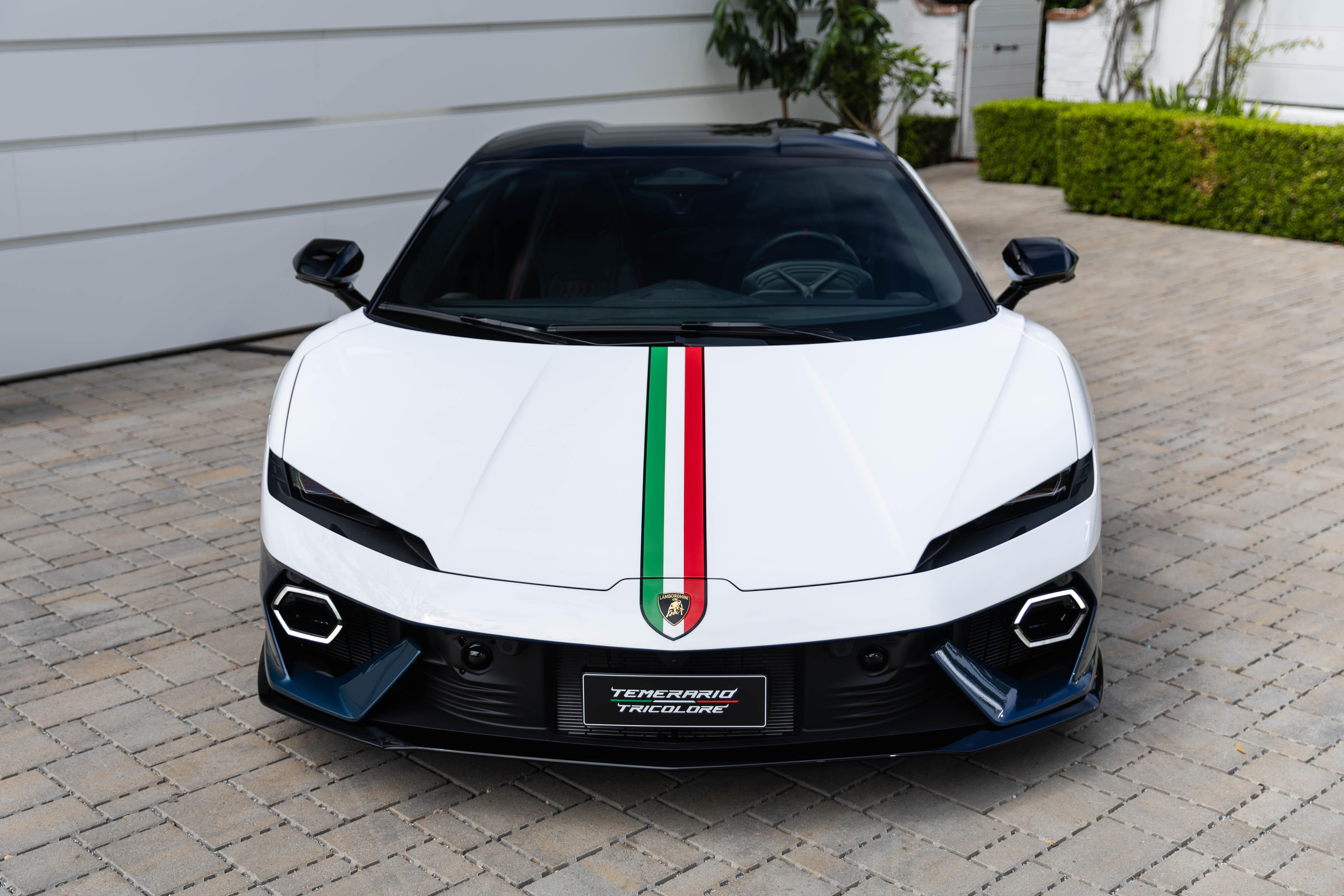 Carro esportivo branco com teto preto, faixas verde, branca e vermelha no capô, estacionado em piso de paralelepípedos. O para-choque tem detalhes azuis e pretos, com a placa TEMERARIO TRICOLORE