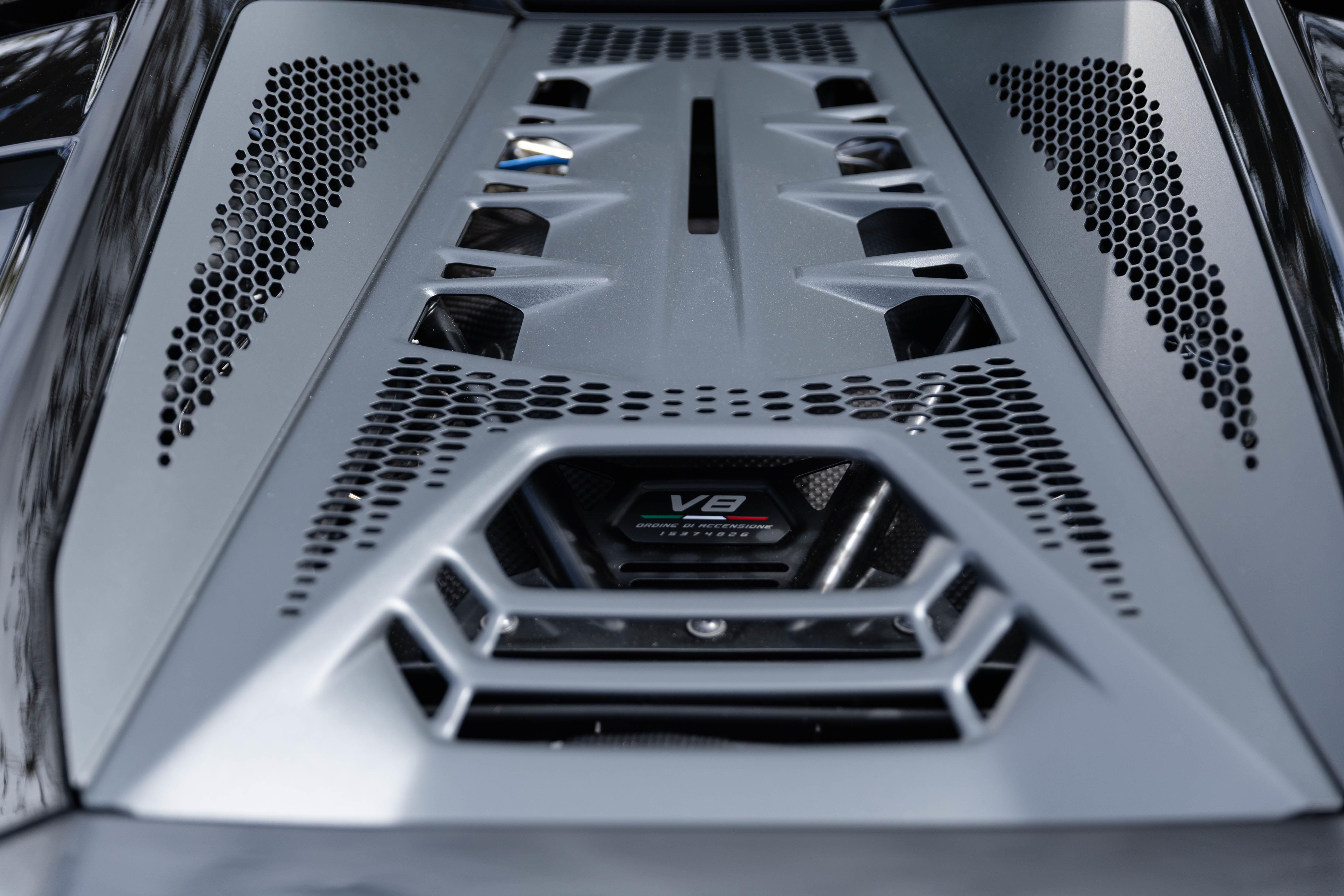 Detalhe do motor V8 de um carro esportivo, com a tampa prateada e aberturas hexagonais para ventilação, exibindo o emblema V8 no centro