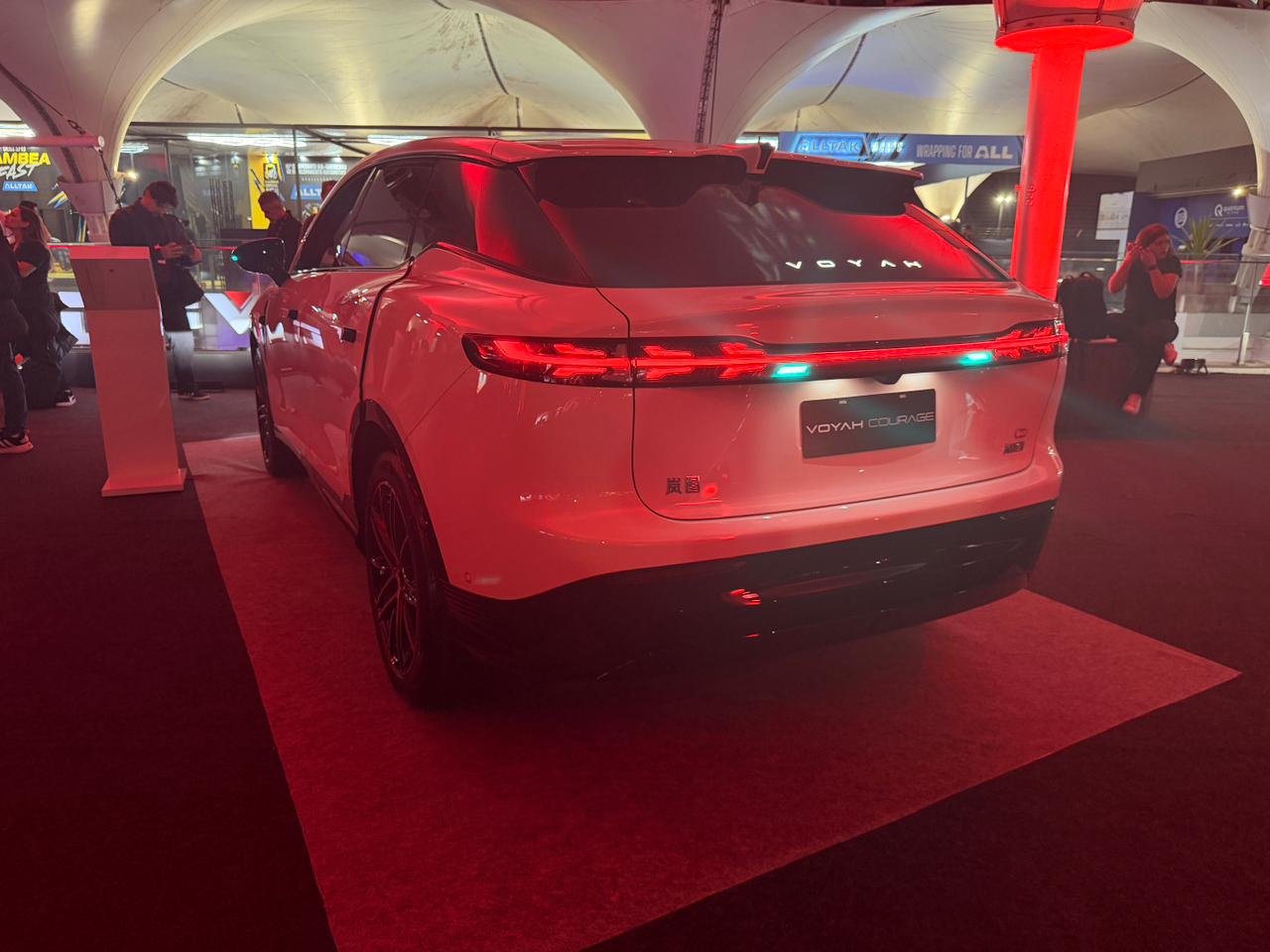 Traseira de um carro SUV branco, modelo Voyah Free, com lanternas vermelhas e uma faixa de luz azul. O carro está em um evento noturno, sob uma tenda branca, com iluminação vermelha no chão e pessoas ao fundo.