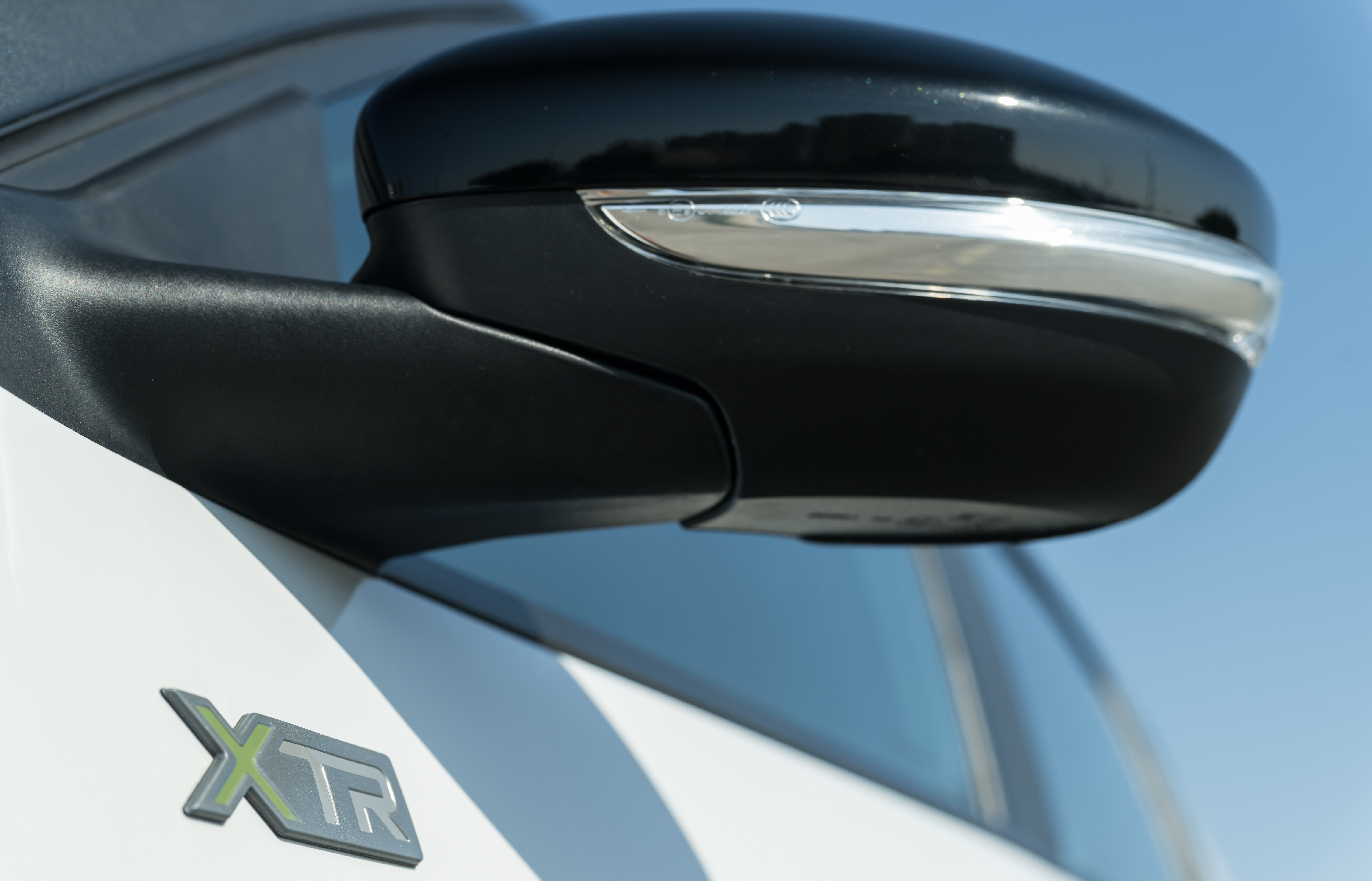 Retrovisor lateral preto de um carro branco, com um detalhe cromado e luz de seta integrada. Abaixo, o emblema XTR em cinza com letras verdes, sob um céu azul claro