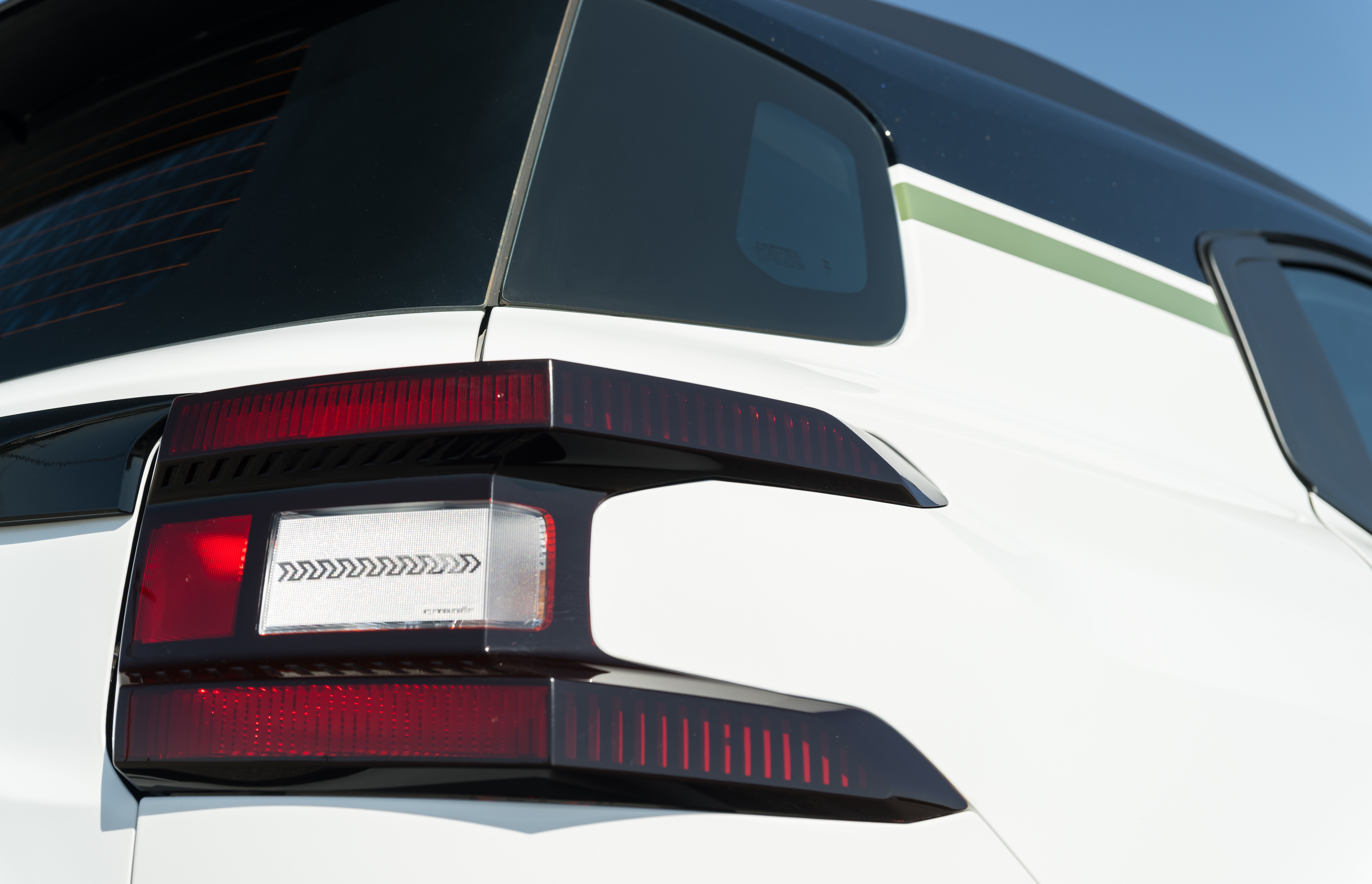 Lanterna traseira direita de um carro branco, com detalhes em preto e uma faixa verde na lateral superior. A lanterna tem formato irregular, com luzes vermelhas e uma área central branca com setas pretas