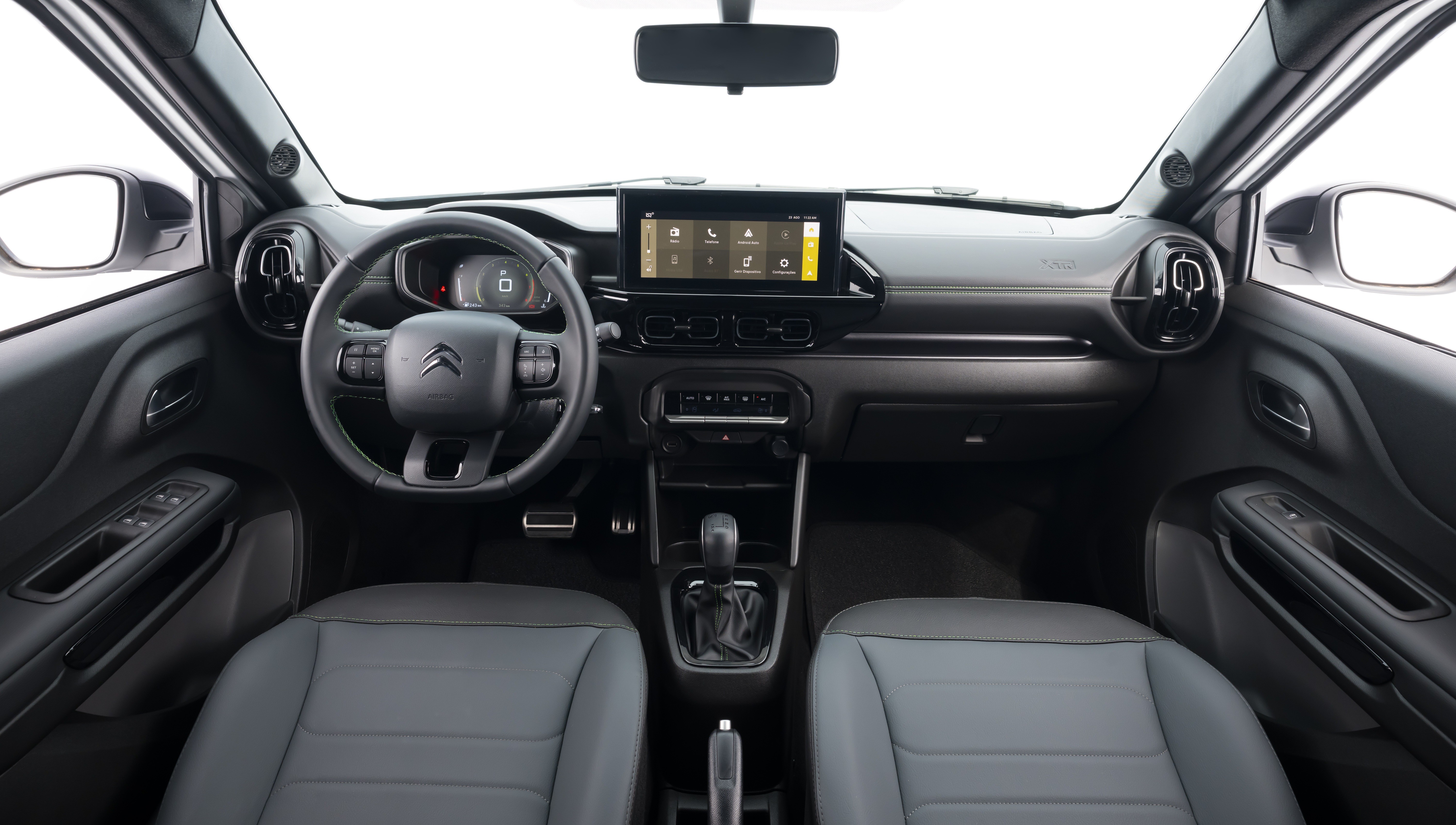 Interior de um carro Citroën C3, com bancos cinza-escuros e painel preto. O volante multifuncional exibe o logo da marca, e uma tela multimídia central mostra ícones de aplicativos. O câmbio automático está no console central, e os espelhos retrovisores laterais são visíveis pelas janelas. O ambiente é moderno e funcional
