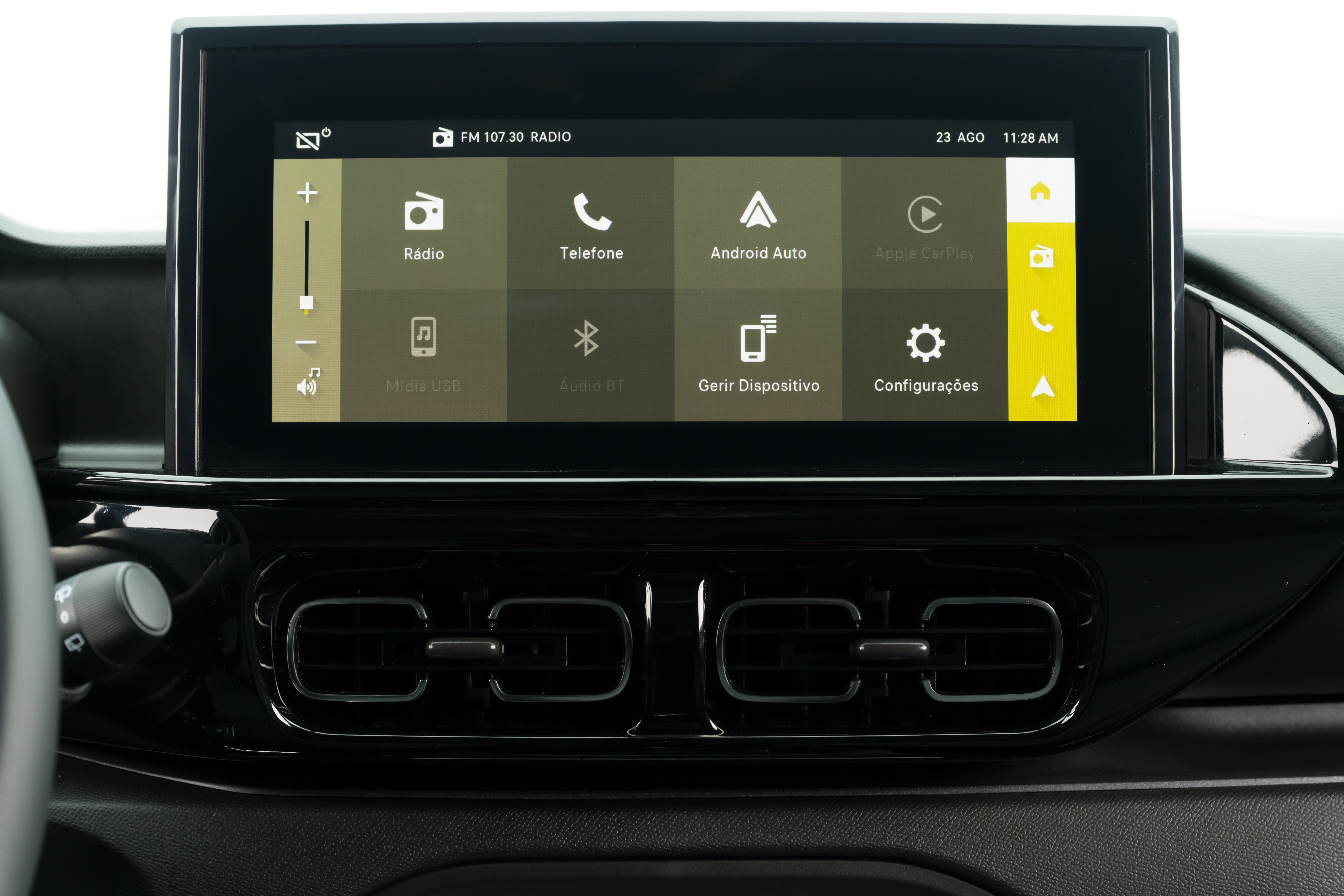 Tela multimídia de carro com menu principal exibindo ícones de Rádio, Telefone, Android Auto, Apple CarPlay, Mídia USB, Áudio BT, Gerir Dispositivo e Configurações. À esquerda, controle de volume e à direita, barra de atalhos em amarelo. Abaixo, duas saídas de ar retangulares no painel preto do veículo