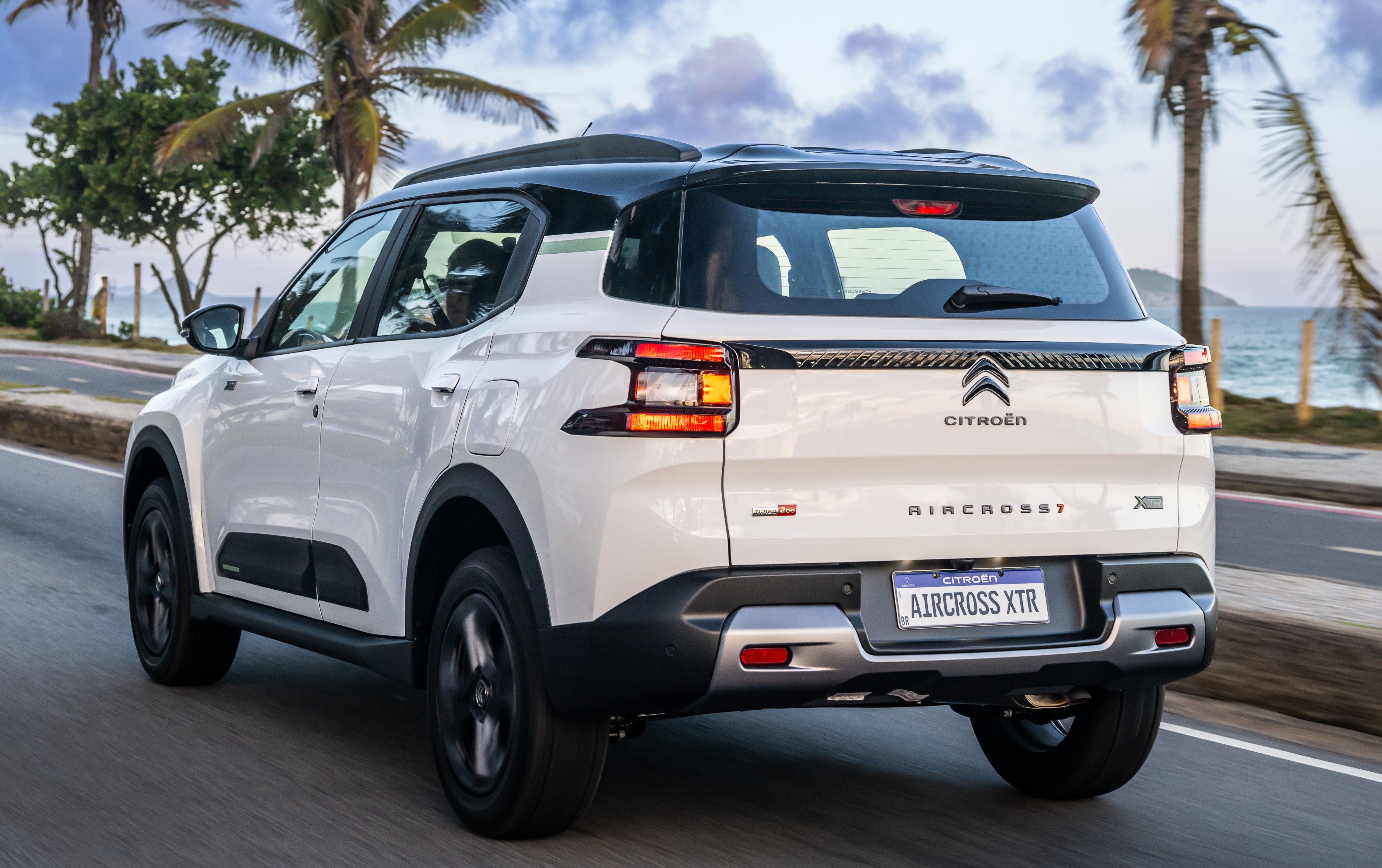 SUV Citroën Aircross XTR branco com teto preto, visto de traseira, em movimento numa estrada costeira com palmeiras e mar ao fundo