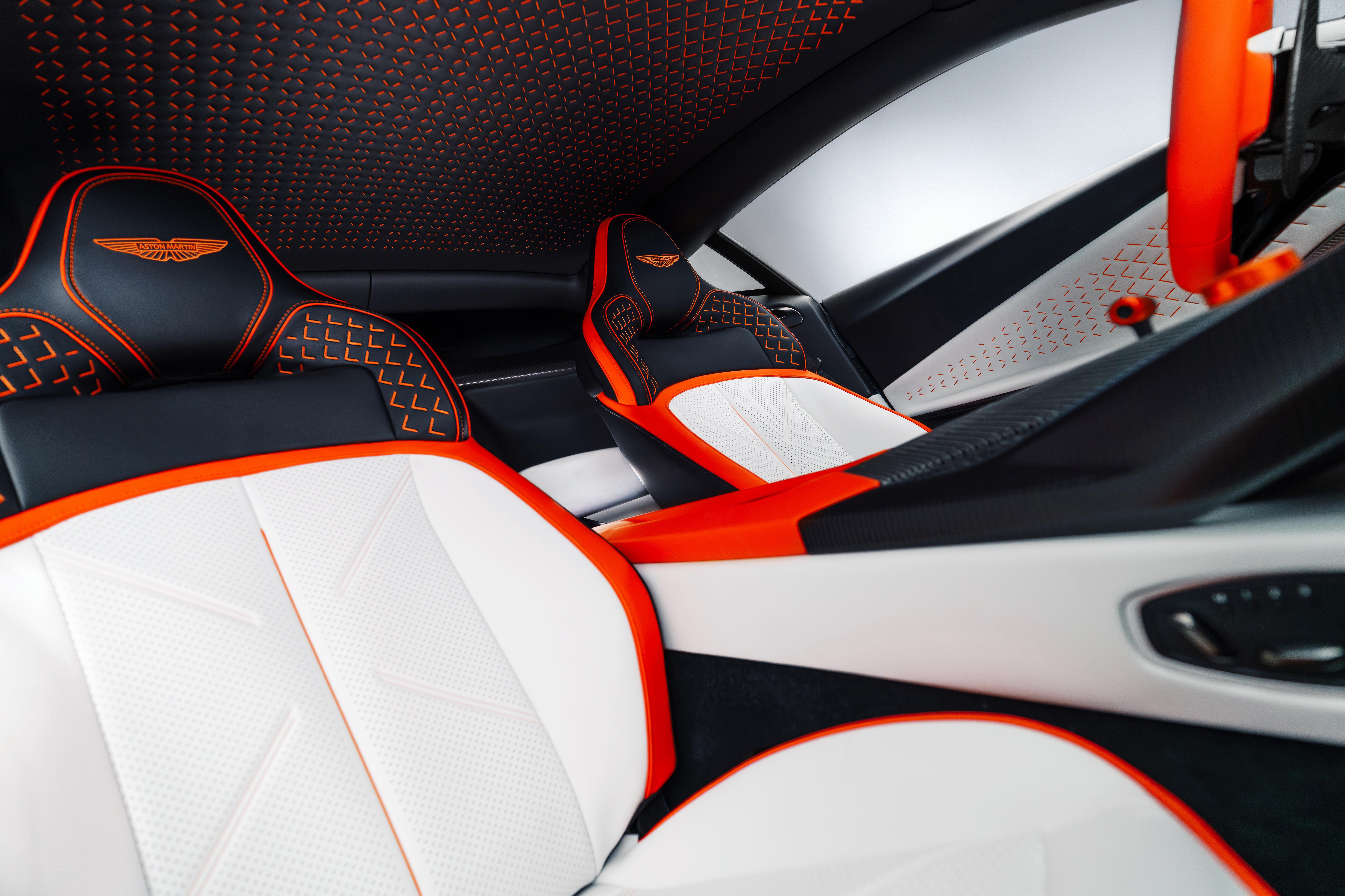 Interior de carro esportivo com bancos pretos e brancos, detalhes em laranja vibrante. O teto e encostos possuem costuras geométricas, e o painel lateral é branco com textura pontilhada