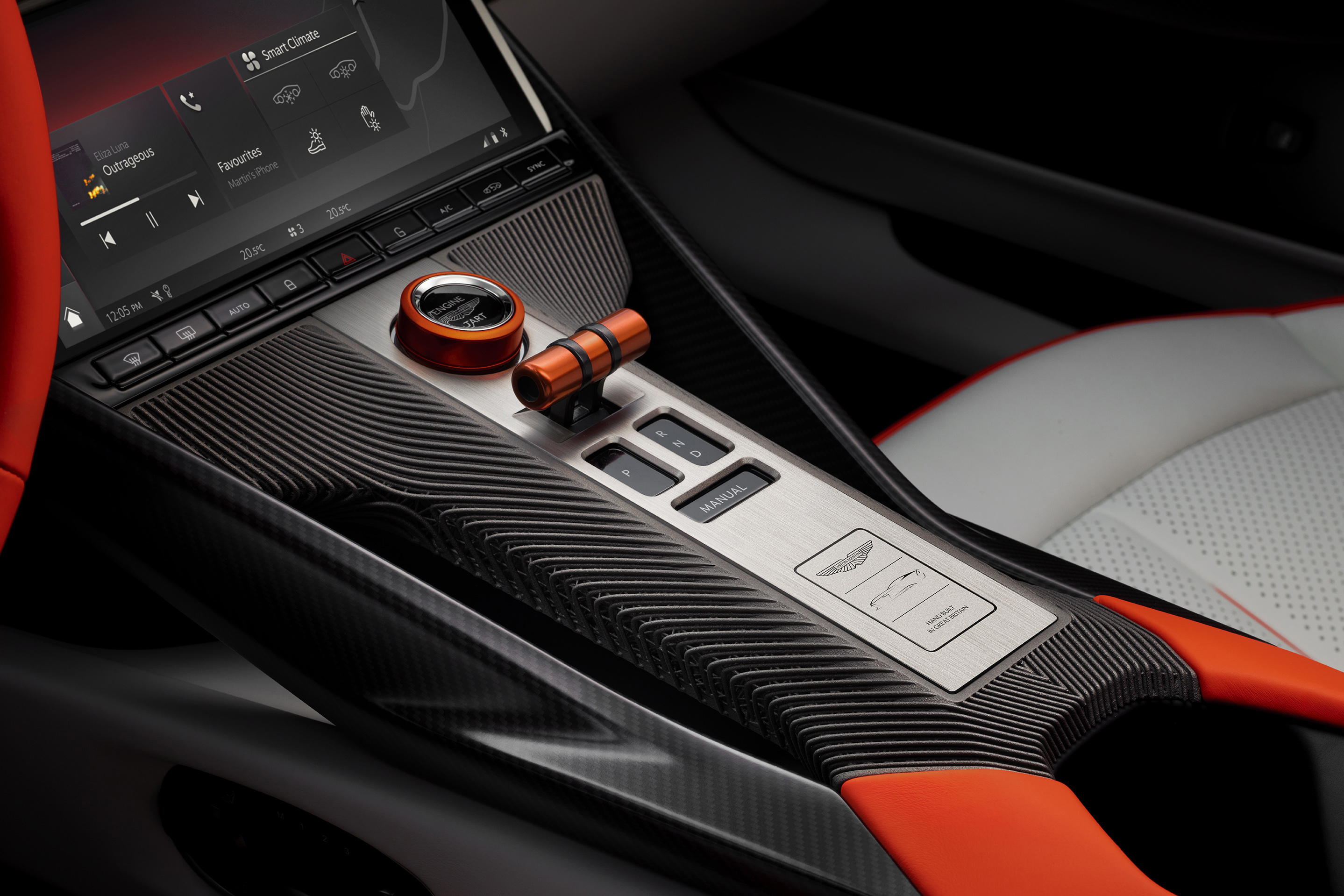 Interior de carro esportivo com console central em fibra de carbono e detalhes laranja. Tela multimídia exibe Smart Climate e Favourites. Botões de controle e um seletor de marcha em formato de alavanca laranja estão visíveis. O banco do passageiro é branco com detalhes vermelhos.