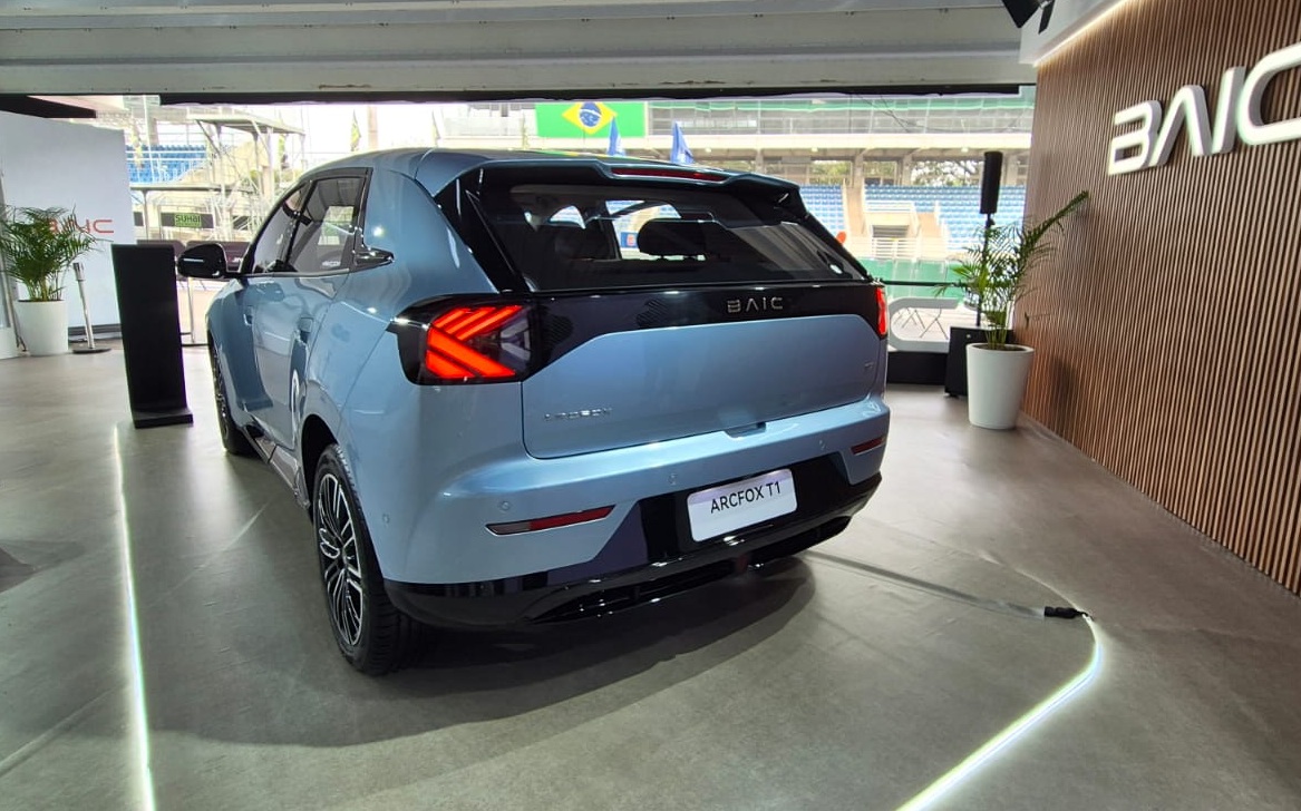 Traseira de um carro SUV azul-claro, modelo Arcfox T1, em um showroom. As lanternas traseiras são vermelhas e o para-choque é preto. Ao fundo, uma parede de madeira com o logo BAIC iluminado e uma bandeira do Brasil.