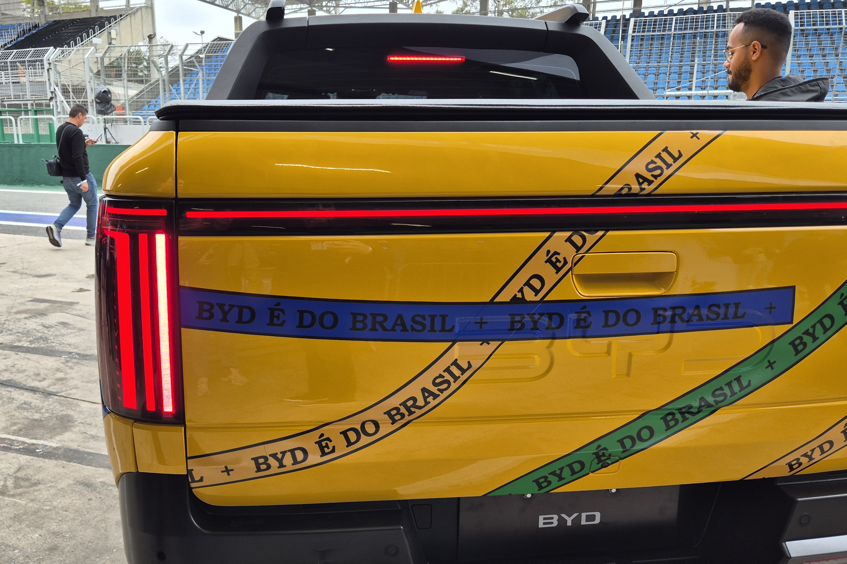 Traseira de uma picape amarela com adesivos BYD É DO BRASIL em faixas azuis, verdes e pretas. Um homem com barba e óculos está no banco do passageiro, e outro homem caminha ao fundo, próximo a uma arquibancada