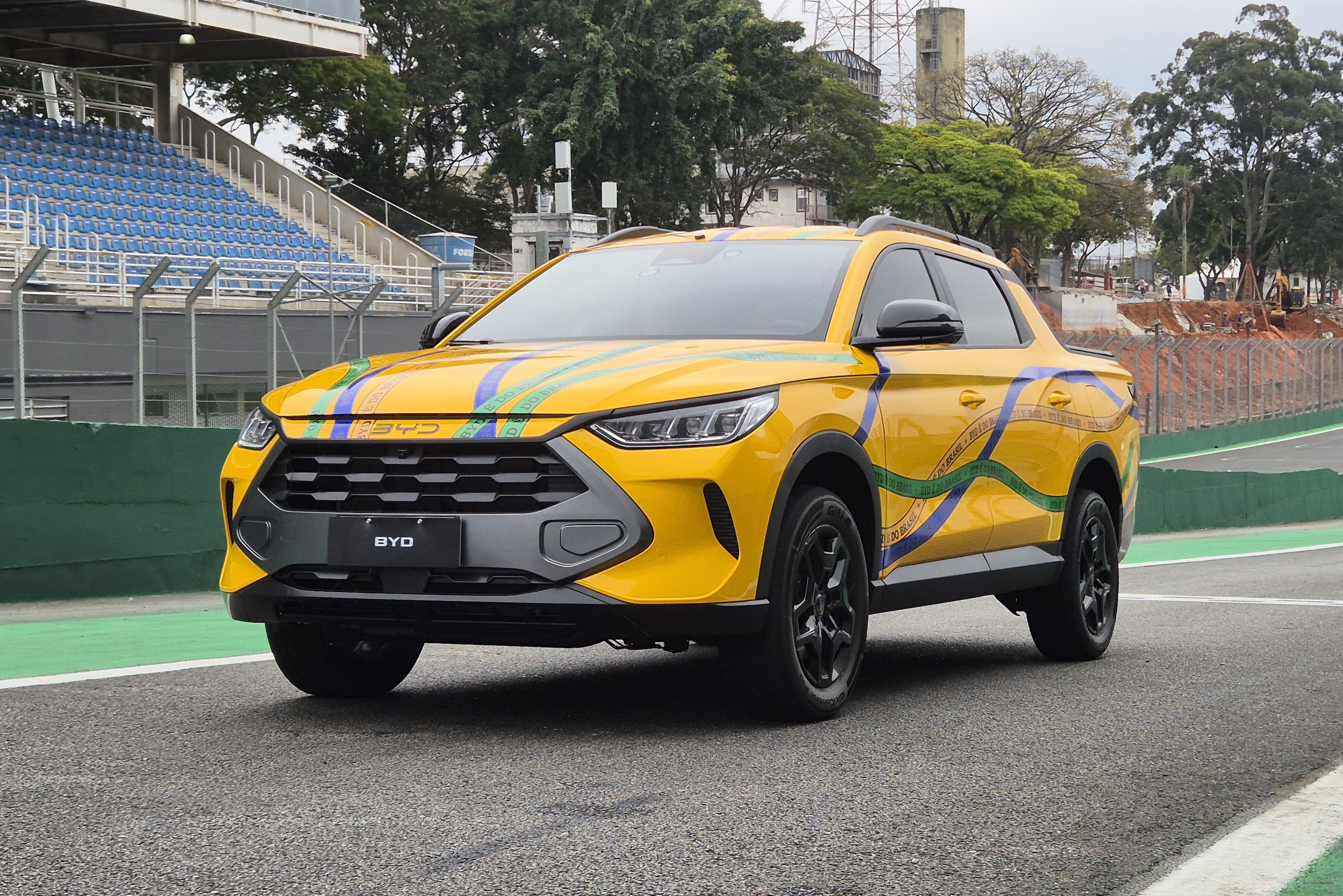 SUV amarelo com detalhes em azul, verde e branco, cores da bandeira brasileira, parado em uma pista de corrida com arquibancadas azuis ao fundo