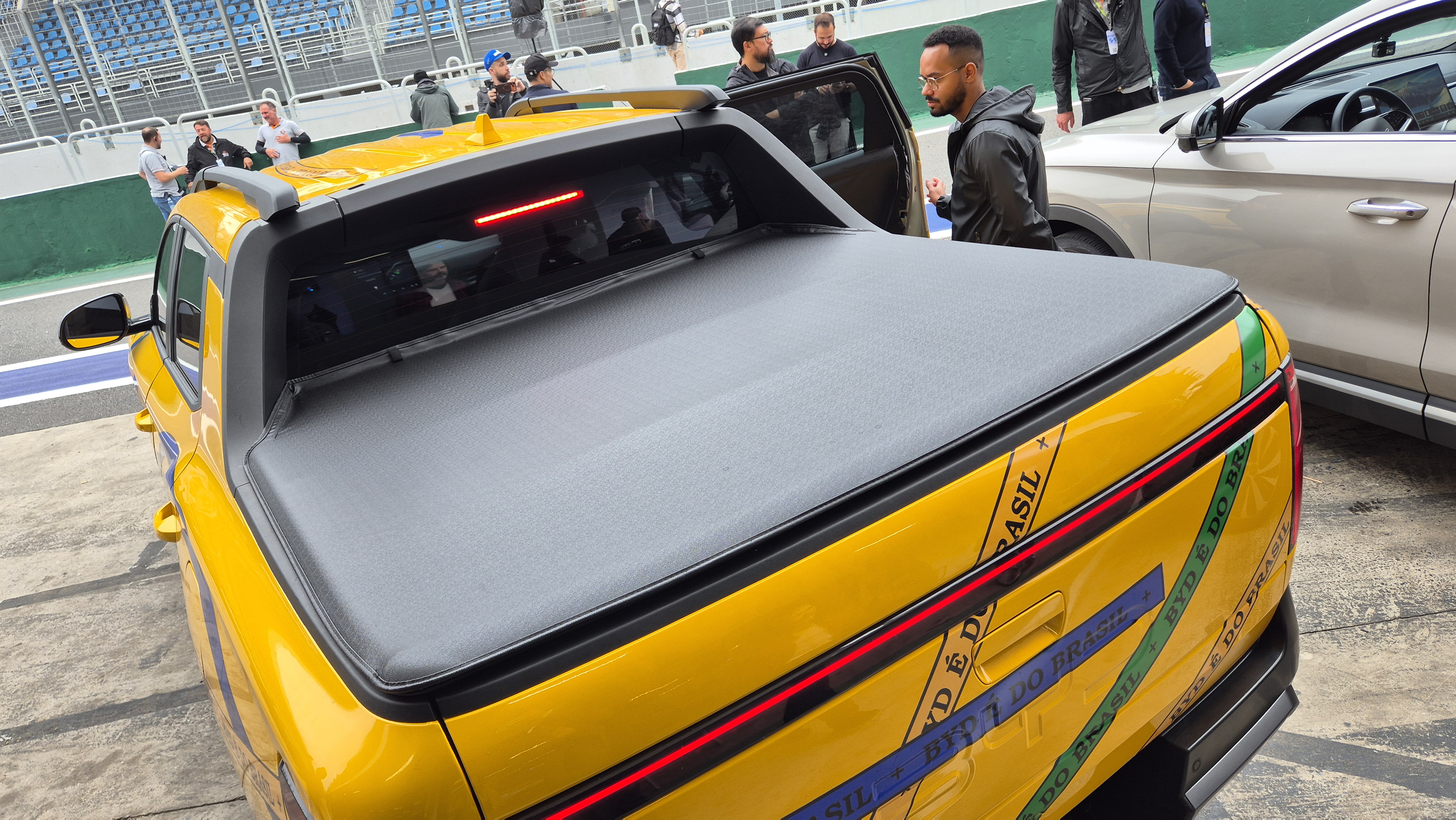 Picape amarela com capota marítima preta, adesivos de fitas do Senhor do Bonfim nas cores da bandeira do Brasil na traseira, em uma pista de corrida. Um homem de óculos e jaqueta preta está ao lado da porta aberta do veículo, enquanto outras pessoas observam ao fundo