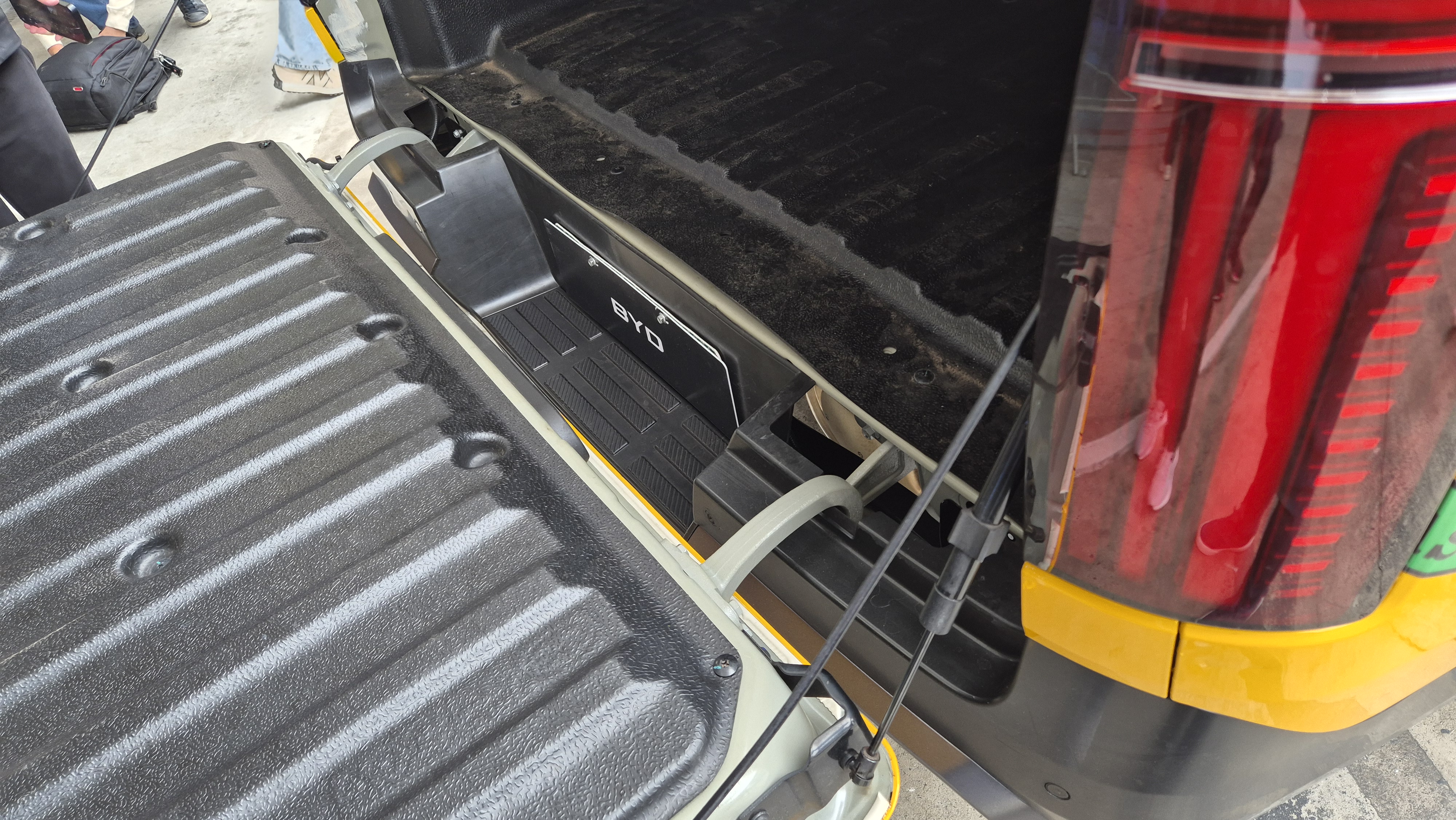 Caçamba de picape com a tampa traseira aberta, revelando um compartimento lateral com o logo BYD e um piso texturizado. A lanterna traseira vermelha do veículo é visível à direita, com reflexos de pessoas.