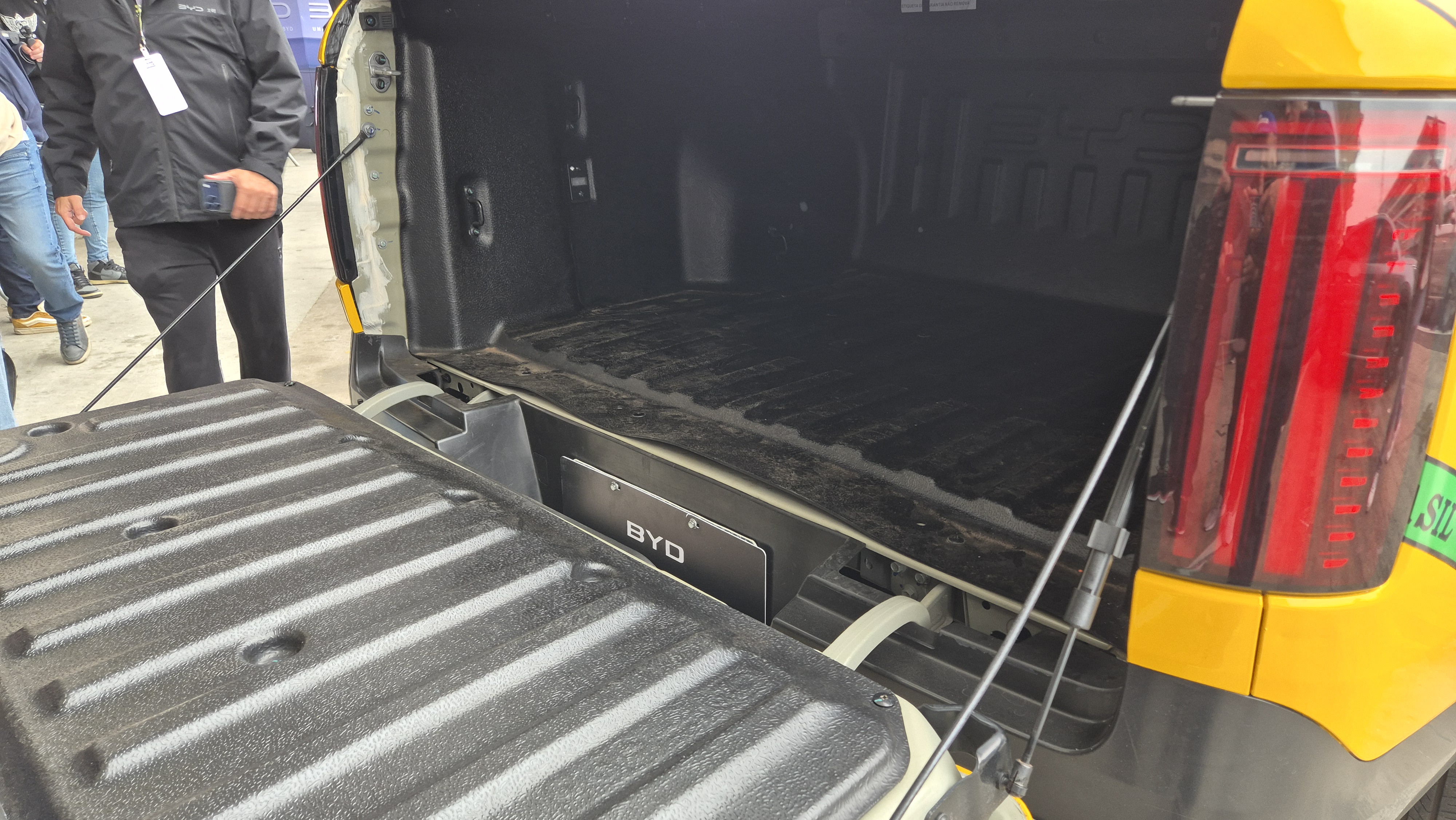 Caçamba de uma picape amarela com a tampa traseira aberta, revelando o interior escuro e texturizado. Uma placa preta com o nome BYD está visível na parte inferior da caçamba. Uma pessoa de jaqueta preta e calça escura está à esquerda, segurando um celular