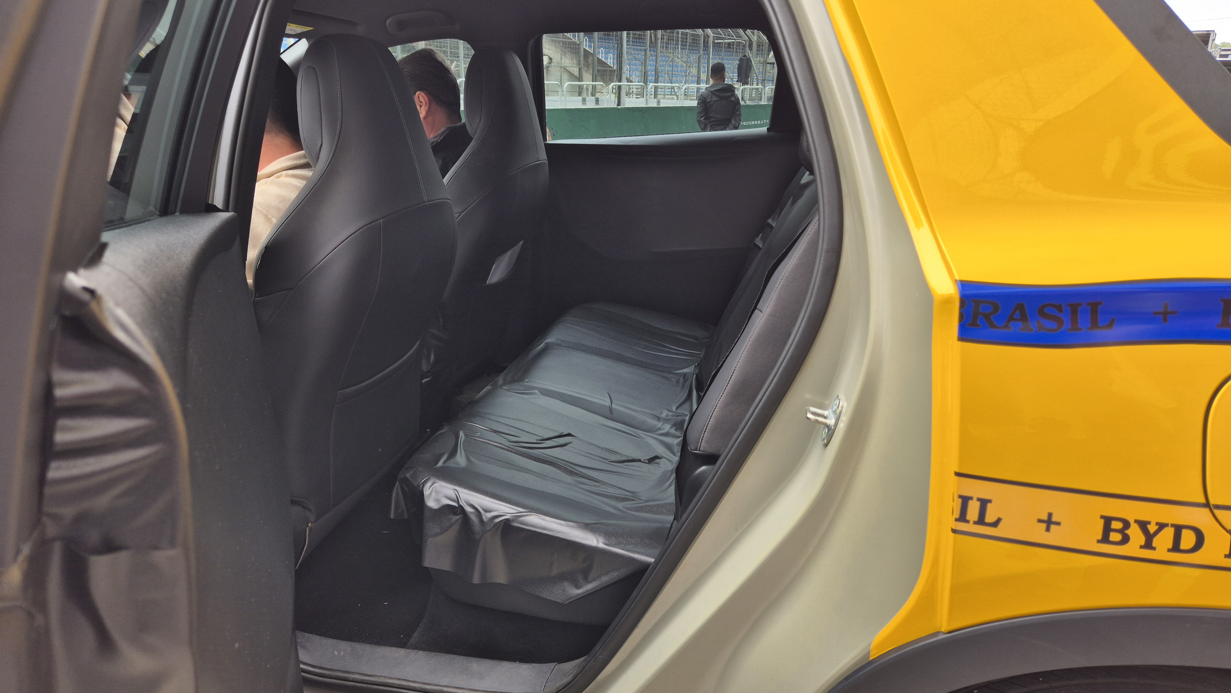 Interior de um carro com bancos pretos, sendo os traseiros cobertos por plástico. Duas pessoas sentadas nos bancos dianteiros, vistas de costas. Pela janela traseira, vê-se uma pessoa em pé e uma área verde. A lateral externa do carro é amarela com uma faixa azul e o texto BRASIL + BYD em preto