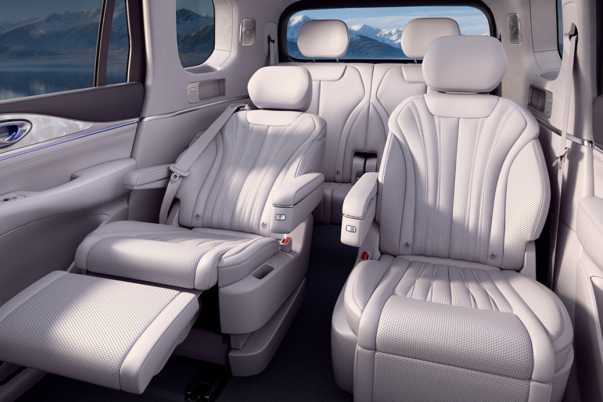Interior de um carro de luxo com bancos de couro branco perfurado, o da esquerda reclinado com apoio para os pés estendido, e o da direita em posição vertical. Ao fundo, montanhas e um lago são visíveis pela janela