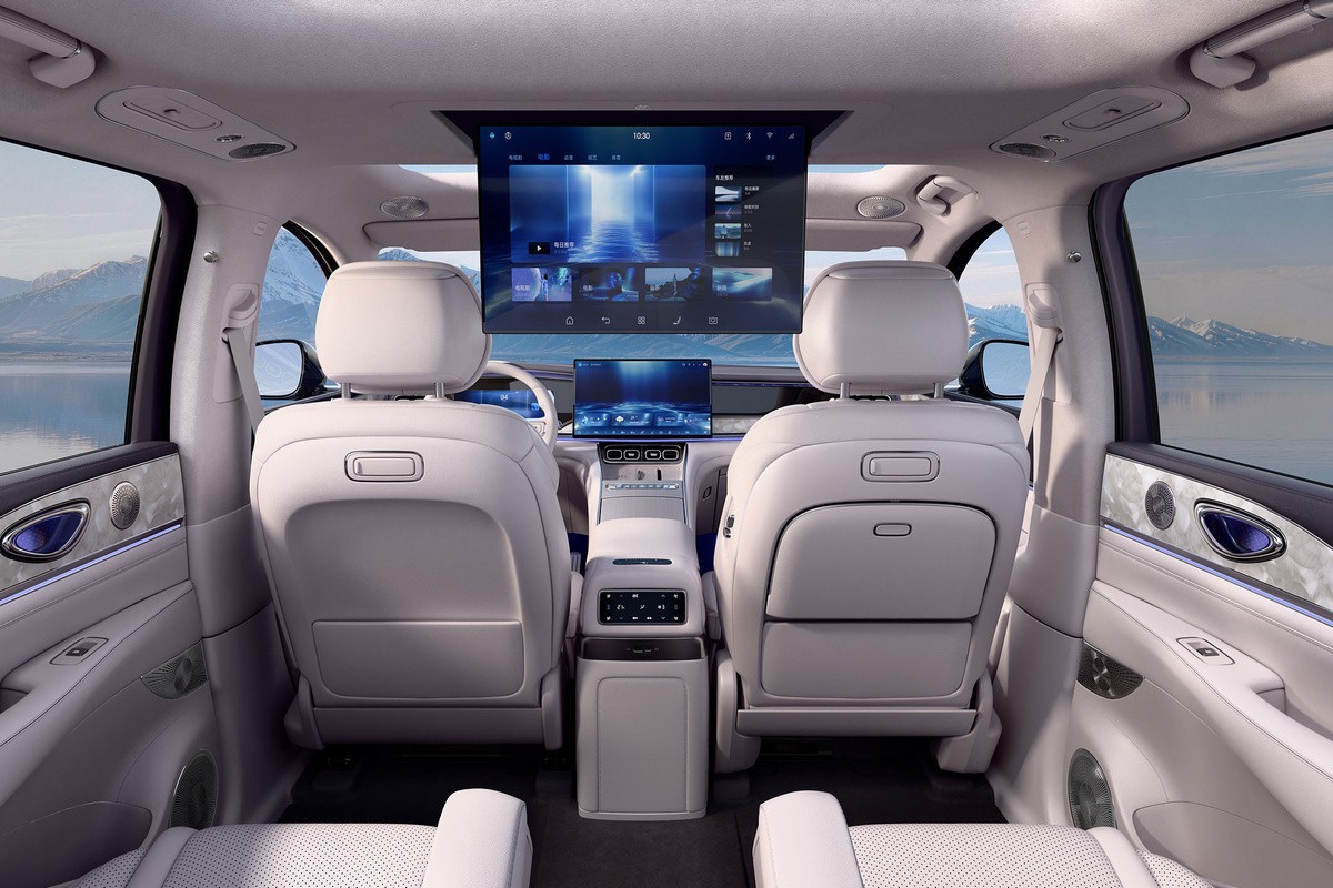 Interior de um carro de luxo com bancos claros, console central e duas telas digitais grandes. Uma tela retrátil no teto exibe um menu de entretenimento, e outra no painel mostra uma paisagem futurista. Ao fundo, montanhas e um lago azul.