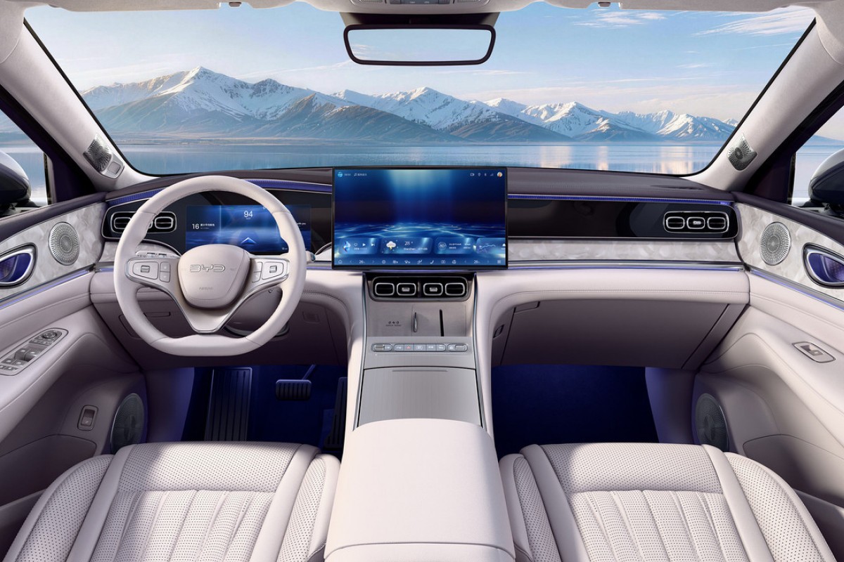 Interior de carro moderno com bancos de couro branco, volante claro e painel com tela grande exibindo uma imagem abstrata azul. Ao fundo, montanhas nevadas e um lago azul sob céu claro, vistos pelo para-brisa