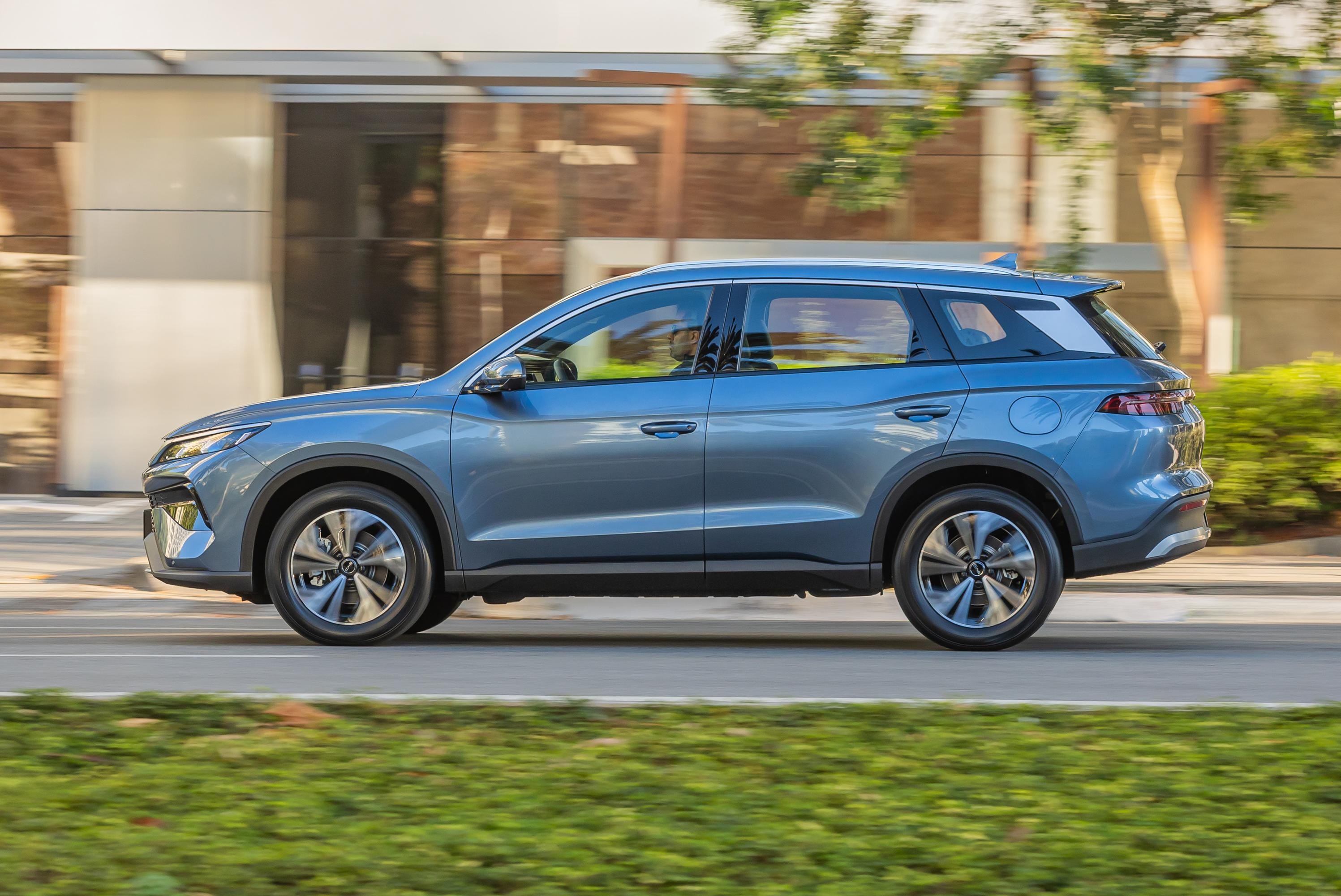 SUV azul-claro em movimento, visto de perfil, com rodas prateadas e detalhes pretos na parte inferior. O fundo desfocado sugere velocidade, com um prédio moderno e árvores verdes