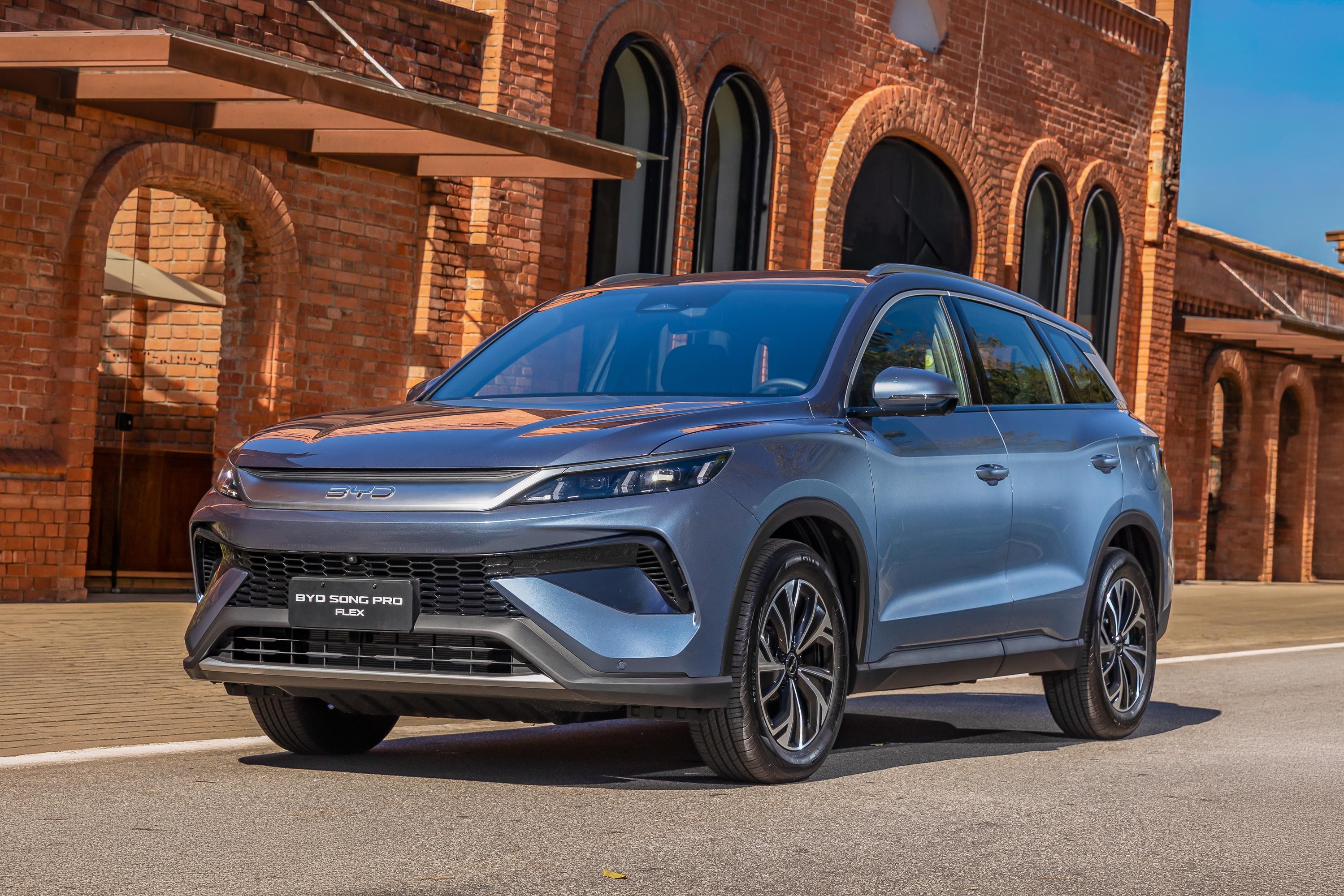 SUV BYD Song Pro Flex azul-claro estacionado em frente a um edifício de tijolos vermelhos com arcos e telhado marrom, sob céu azul