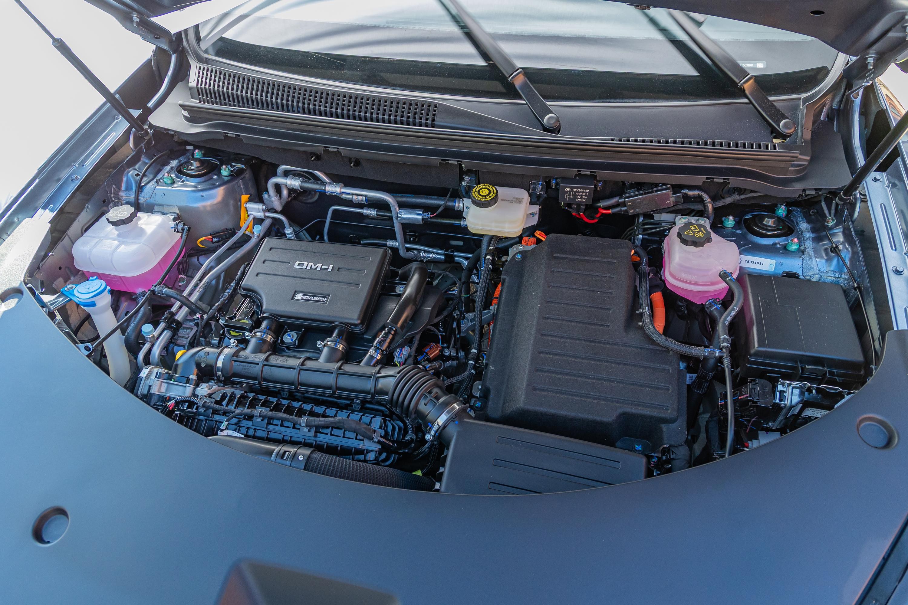 Motor de carro híbrido com capô aberto, mostrando componentes como reservatórios de fluidos rosa e branco, cabos e caixas pretas do sistema elétrico, e o motor a combustão com a inscrição DM-i