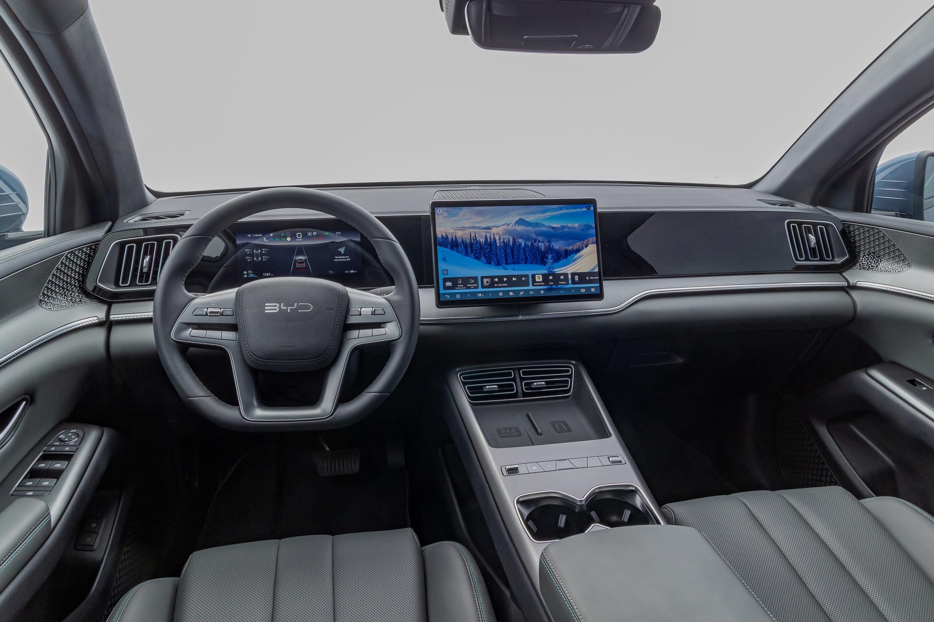 Interior de um carro moderno com volante multifuncional, painel digital e uma tela central grande exibindo uma paisagem de montanhas nevadas. O console central possui porta-copos e carregadores sem fio, e os bancos são de couro cinza escuro