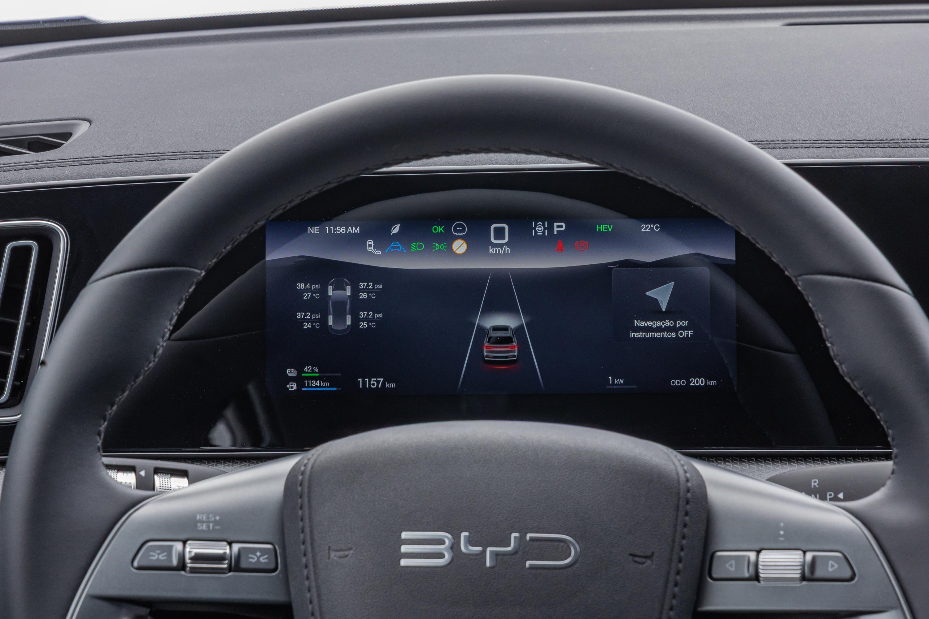 Painel digital de um carro BYD com volante em primeiro plano. O painel exibe informações como hora, temperatura, pressão dos pneus, nível de bateria, autonomia e um carro vermelho em uma estrada, indicando o sistema de assistência ao motorista