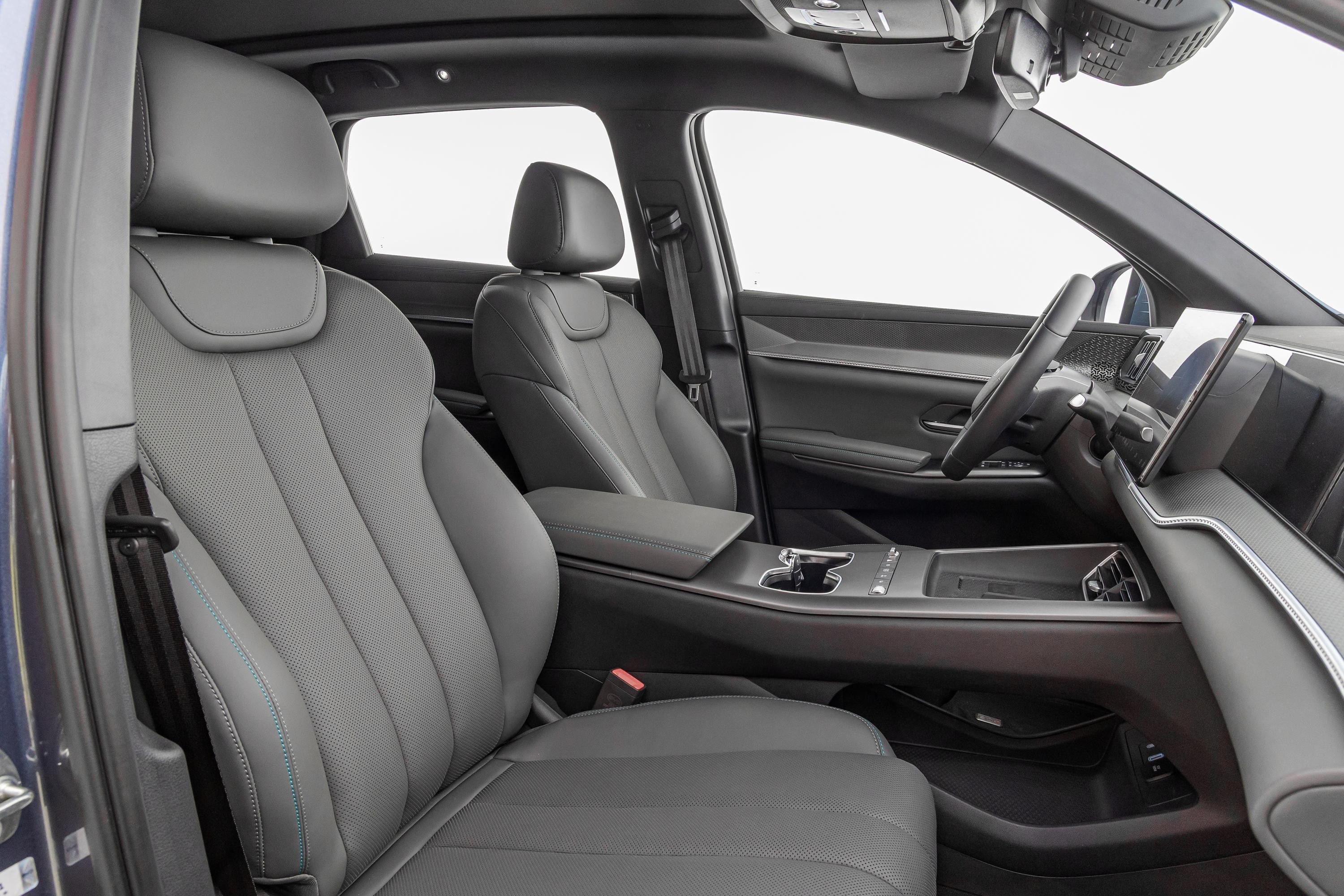 Interior de um carro moderno com bancos de couro cinza-escuro e costuras azuis, console central com porta-copos e tela multimídia inclinada. O volante escuro e o painel minimalista são visíveis, com o teto solar panorâmico ao fundo. A luz natural entra pelas janelas, destacando o design elegante do veículo