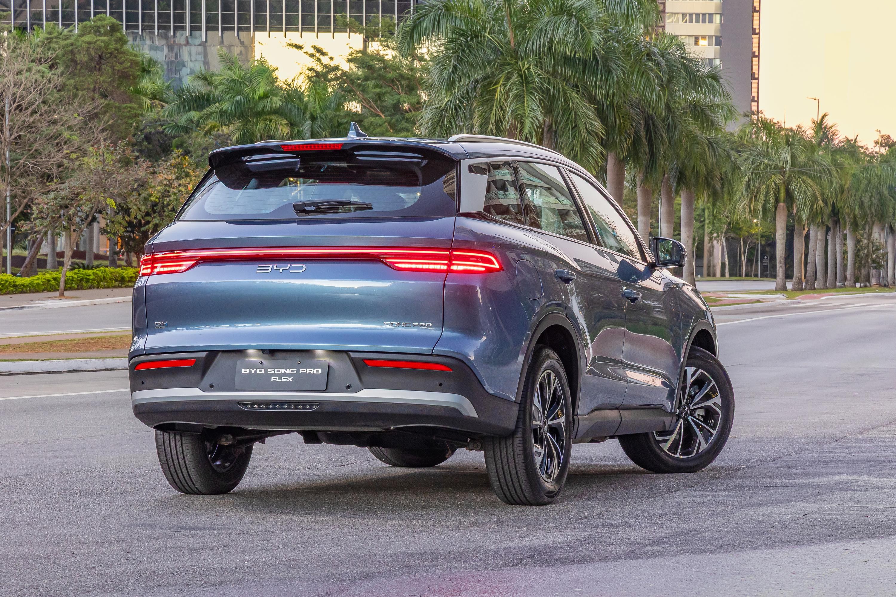 SUV BYD Song Pro Flex azul-escuro, visto de traseira e lateral, parado em rua asfaltada. As lanternas traseiras estão acesas, e o para-choque tem detalhes cinzas. Ao fundo, palmeiras e prédios modernos sob céu claro