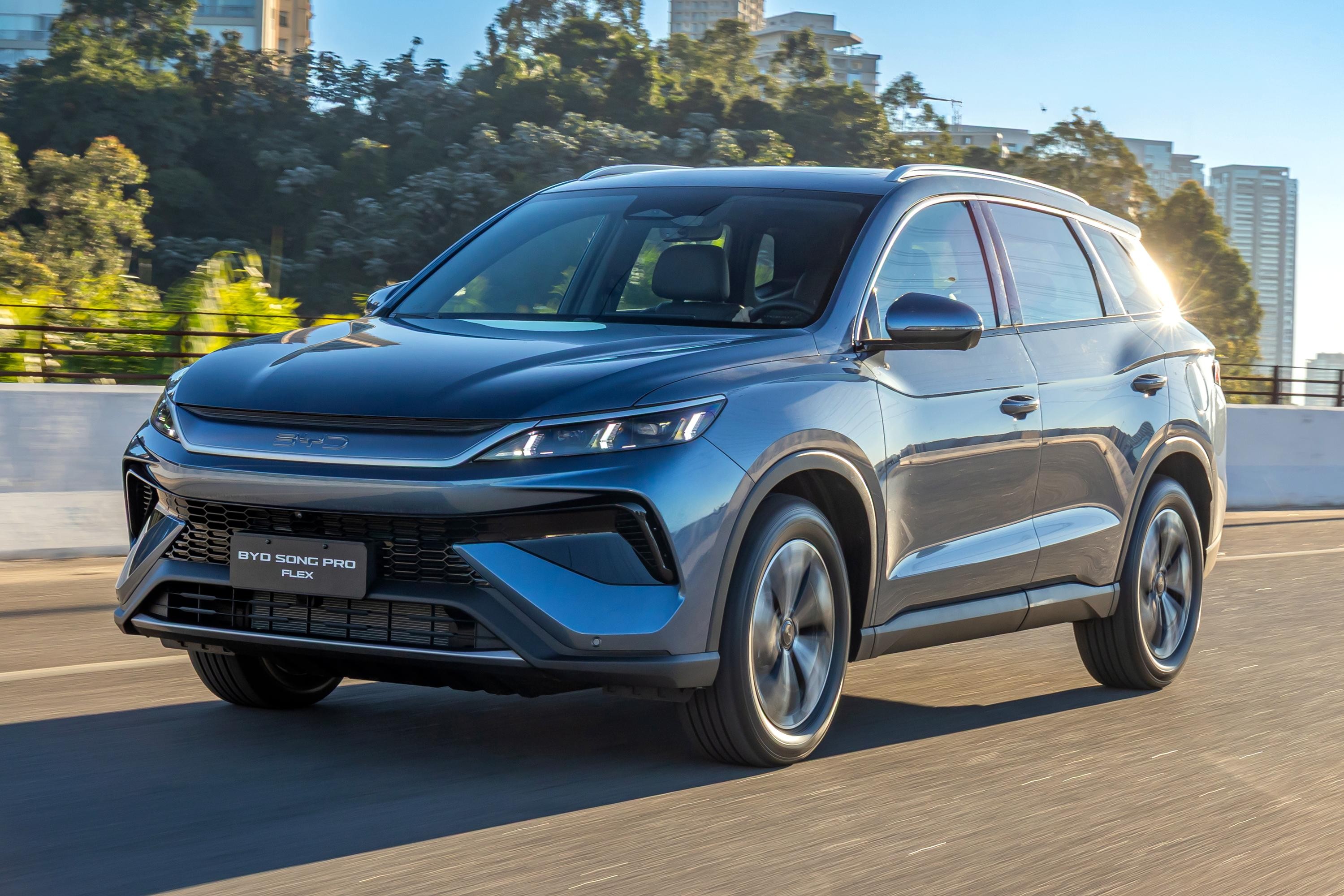 SUV BYD Song Pro Flex azul-escuro em movimento, visto de frente e lateral, com rodas girando. O carro está em uma estrada asfaltada, com árvores e prédios ao fundo sob um céu azul claro. O sol reflete na lateral direita do veículo