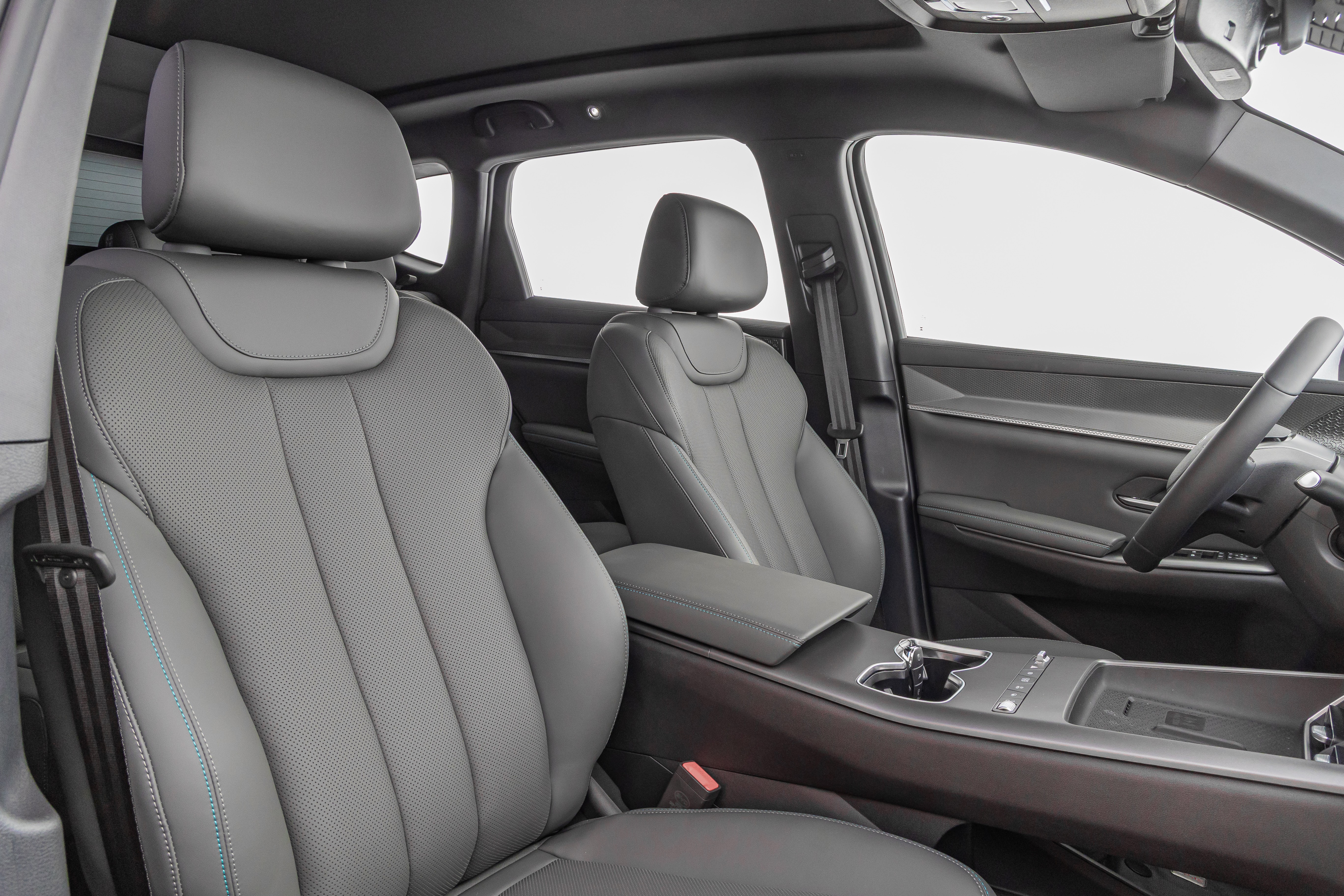 Interior de um carro moderno com bancos de couro cinza perfurado e costura azul. O banco do motorista está em primeiro plano, com o banco do passageiro e o console central visíveis ao fundo. O volante e parte do painel também aparecem à direita