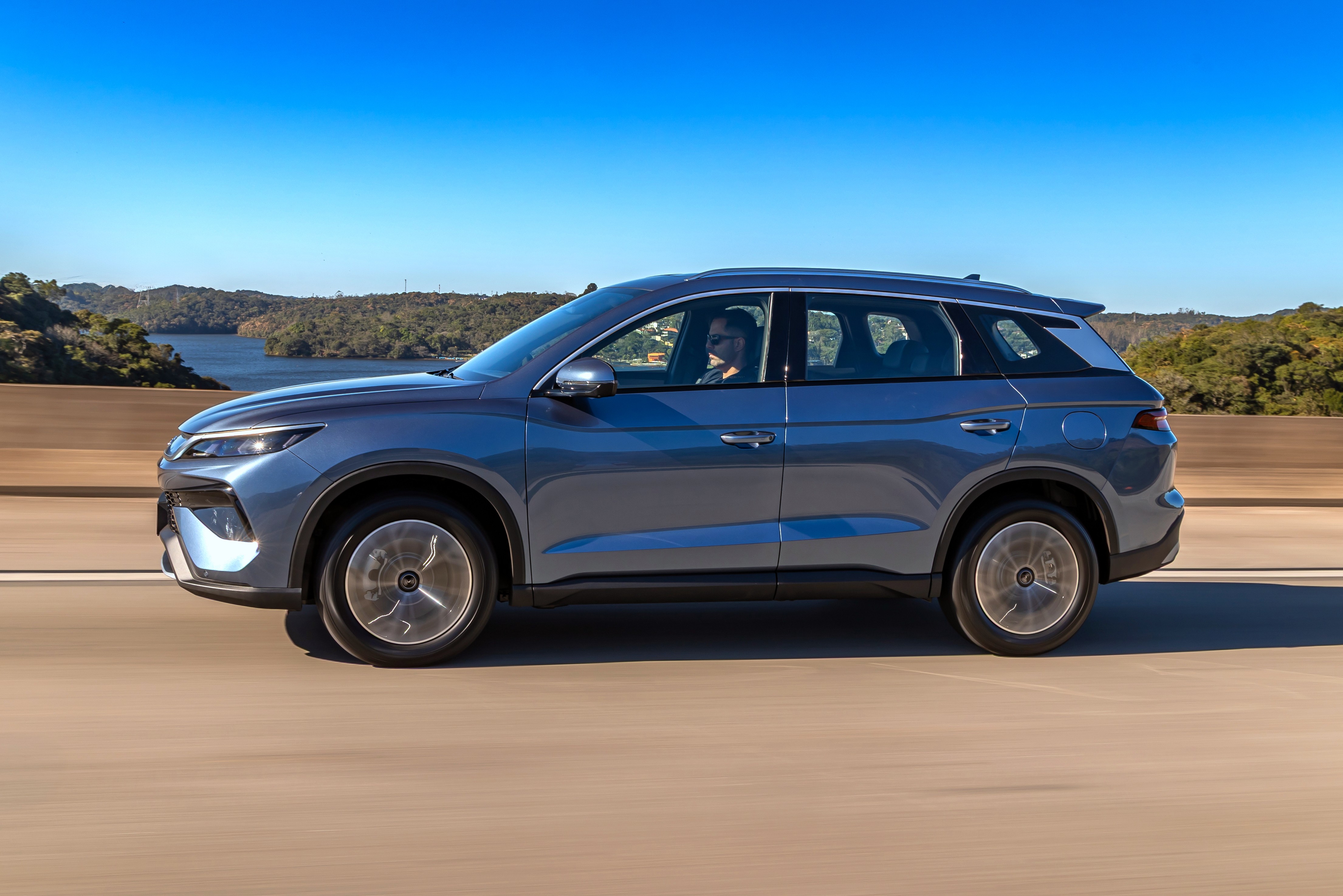 SUV azul-acinzentado em movimento na estrada, com um motorista visível. Ao fundo, um lago cercado por vegetação e um céu azul claro