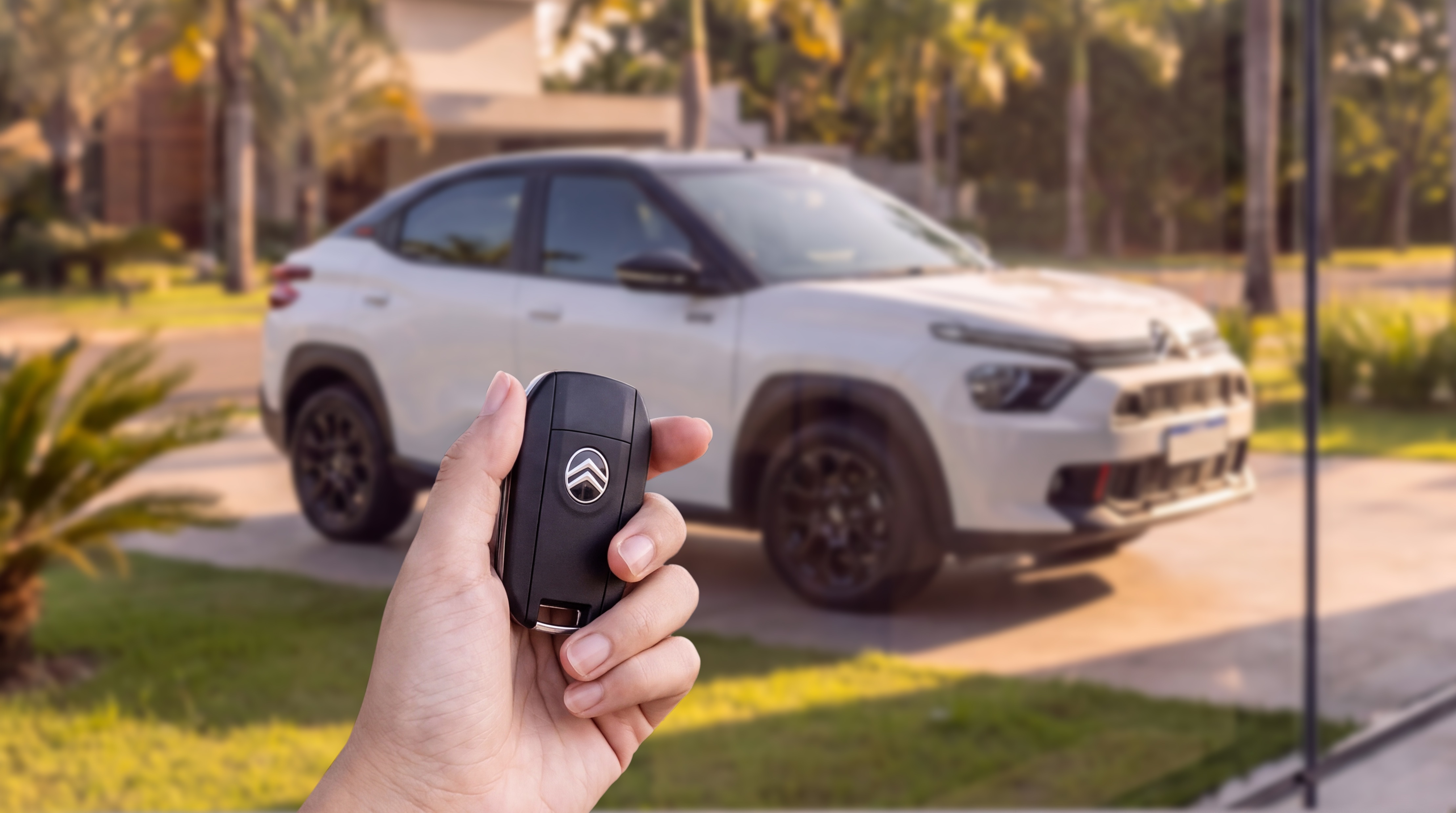 Mão segurando uma chave de carro preta com o logotipo da Citroën, com um carro branco e preto desfocado ao fundo, estacionado em uma garagem com gramado e palmeiras