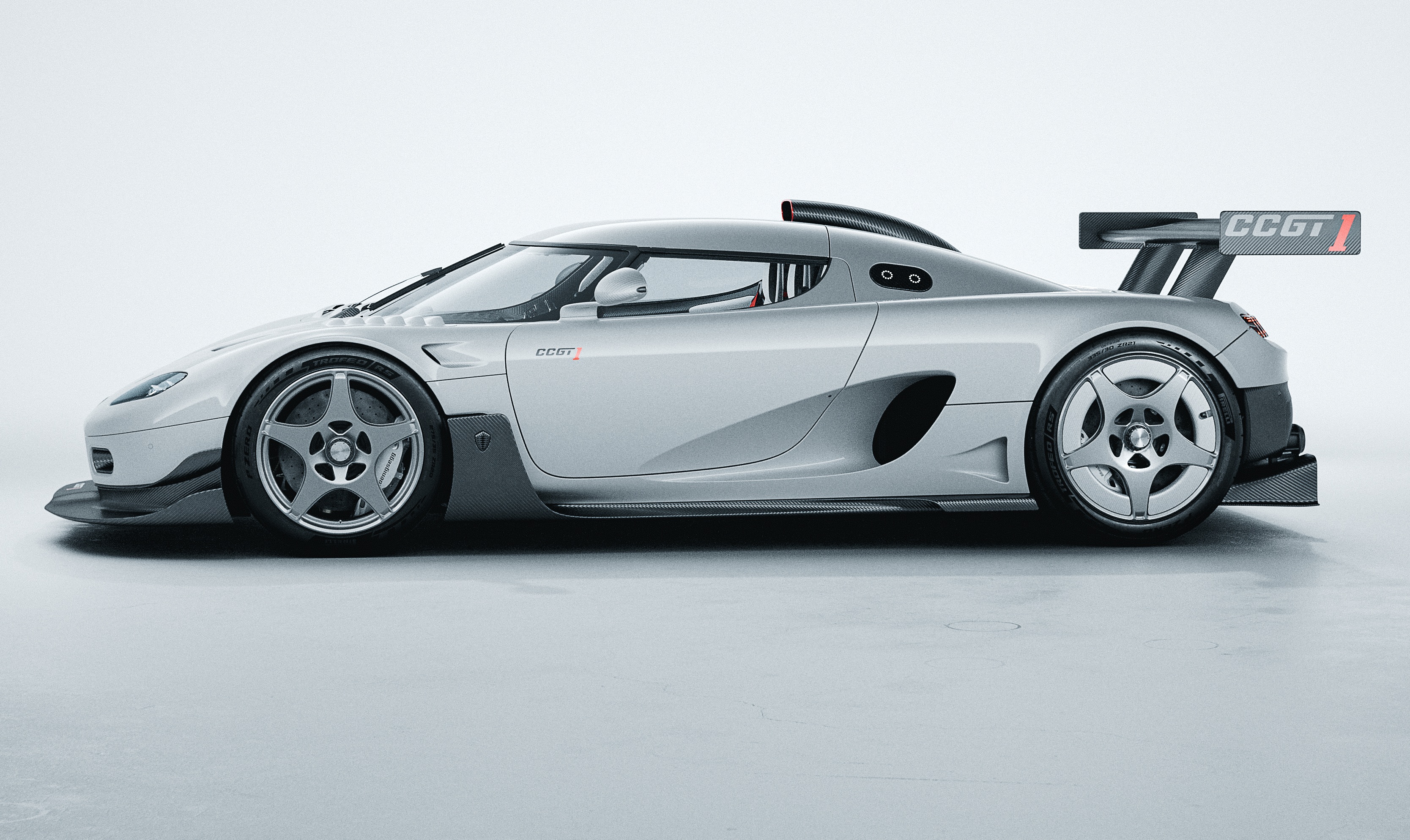 Carro esportivo Koenigsegg CCGT prateado, visto de lado, com aerofólio traseiro e rodas de cinco raios, em um estúdio branco